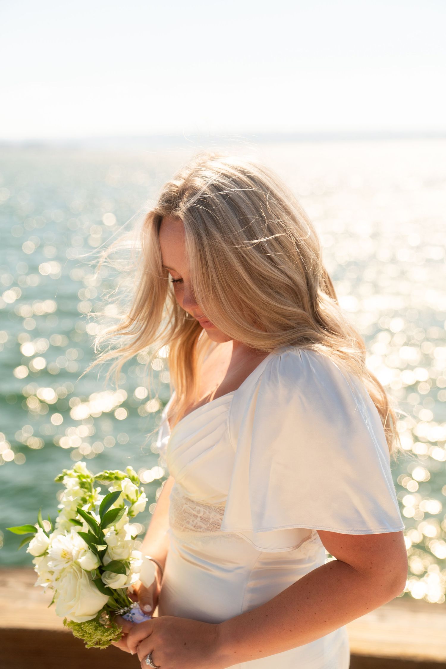 A woman in a white dress holds a bouquet against sparkling water at sunset, creating a romantic coastal scene.