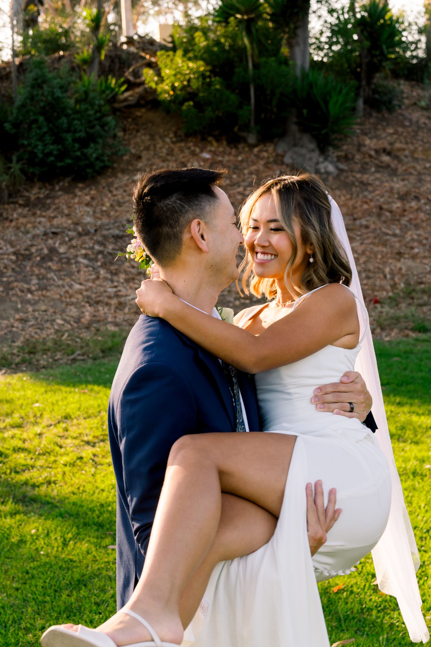 A couple shares a joyful moment in their wedding attire outdoors on a sunlit lawn surrounded by greenery.