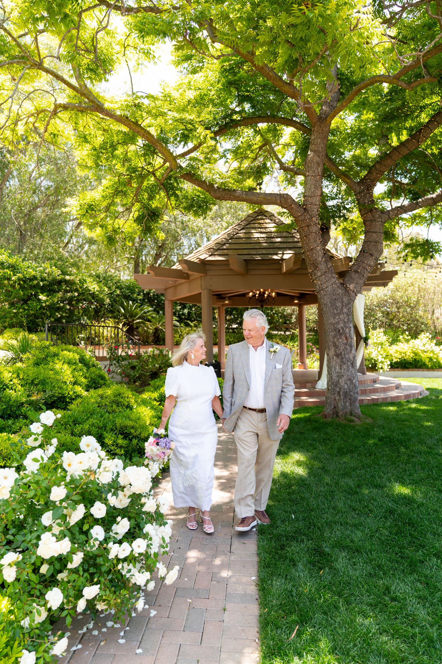 A couple strolls along a brick path in a lush garden with blooming white flowers and mature trees overhead.