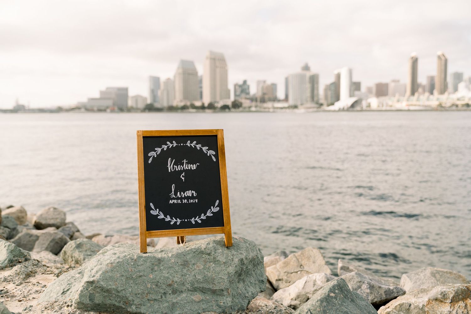 A chalkboard sign stands on rocky shoreline with San Diego skyline in the background.