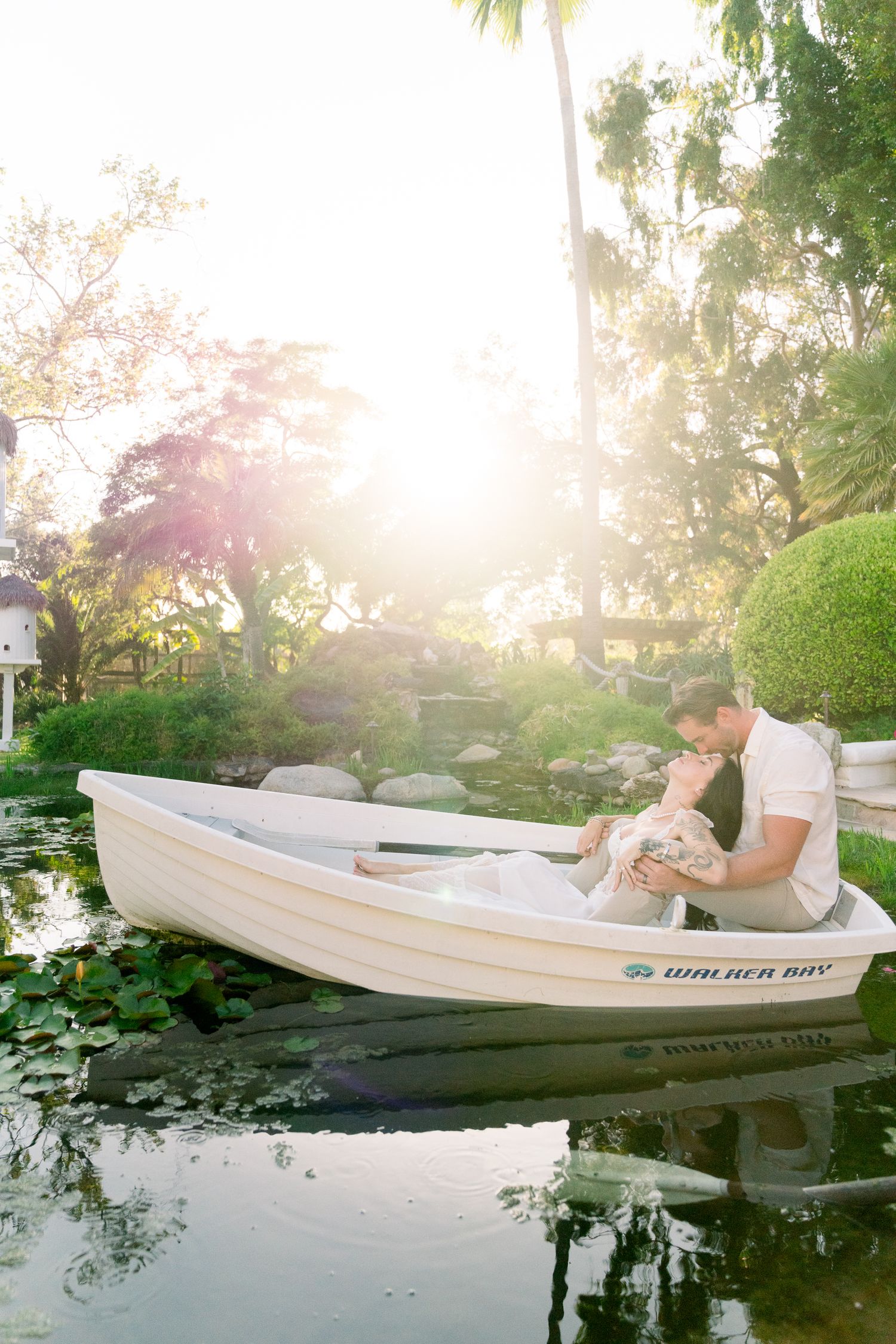 Romantic couple sitting in a white rowboat on a tranquil pond during golden hour with palm trees and lush greenery.