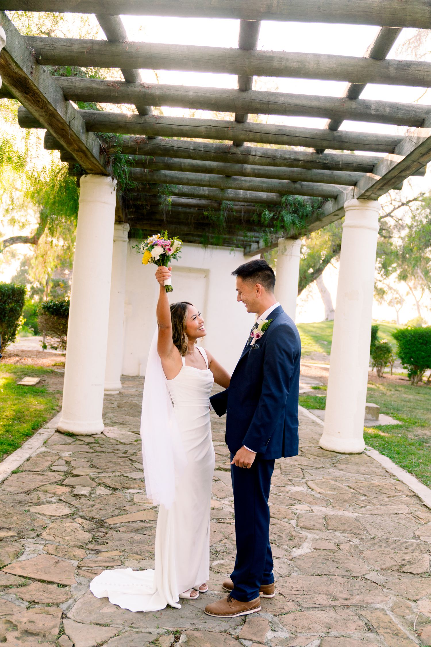 A joyful wedding couple celebrates under a columned pergola with stone flooring in an outdoor garden setting.