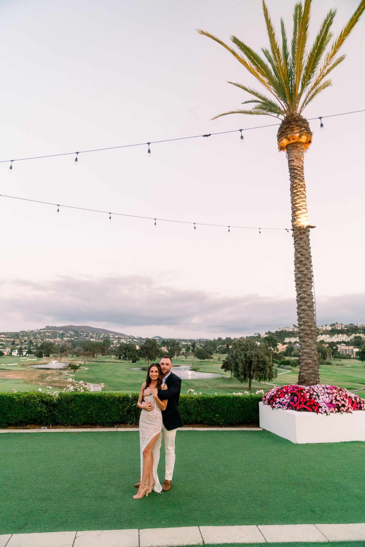 Elegant couple dancing on a terrace with palm trees and string lights at sunset.