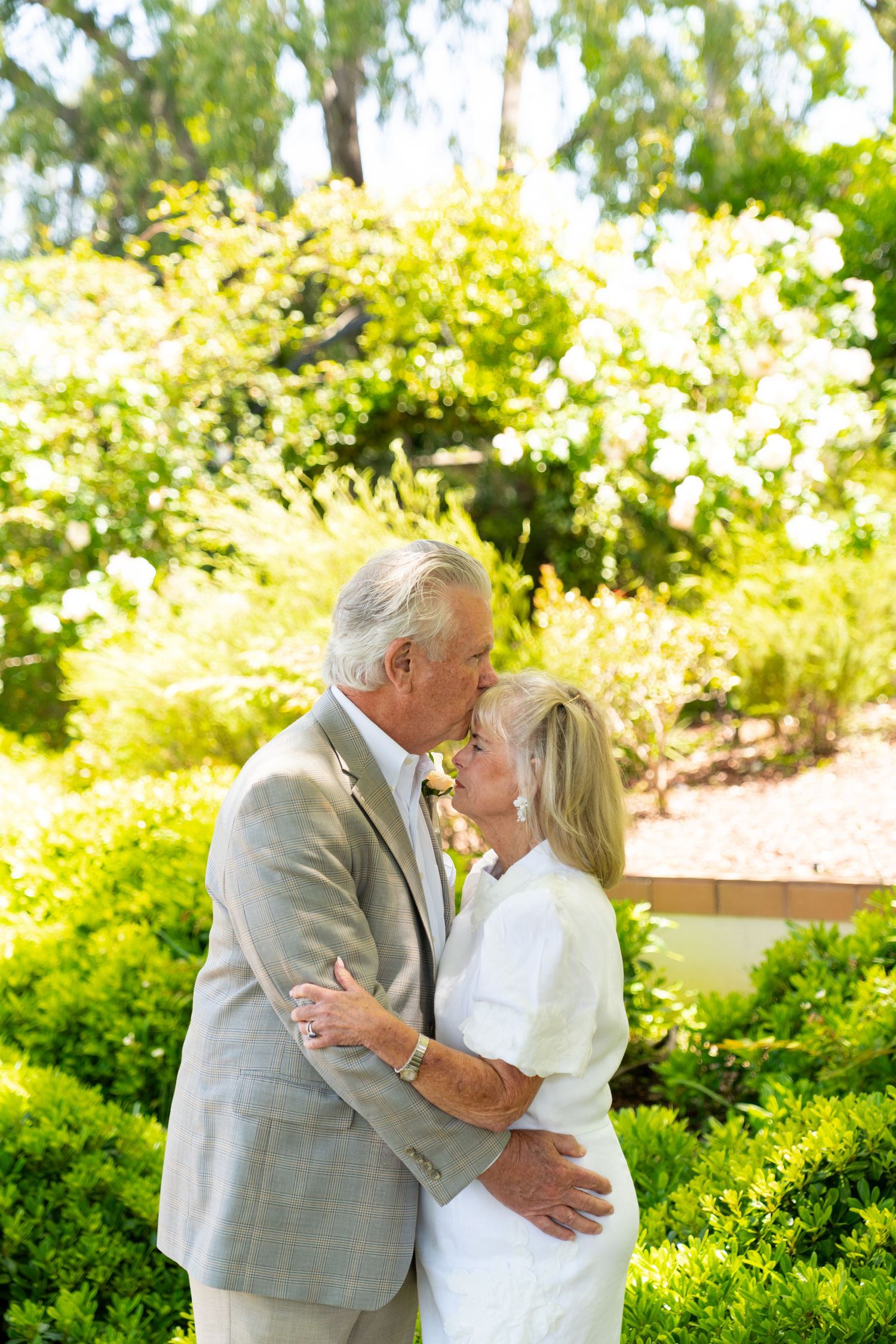 Senior couple embraces lovingly in a sunlit garden with lush green foliage and bright summer light.