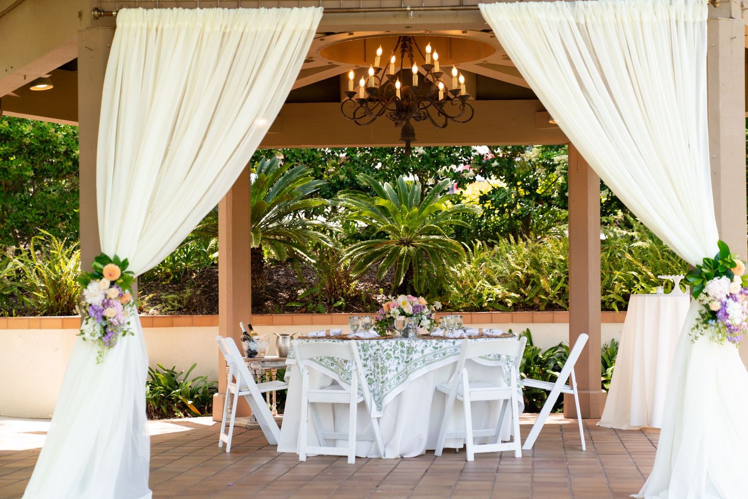 An elegant outdoor wedding gazebo decorated with white drapes, chandelier, dining tables and floral arrangements.