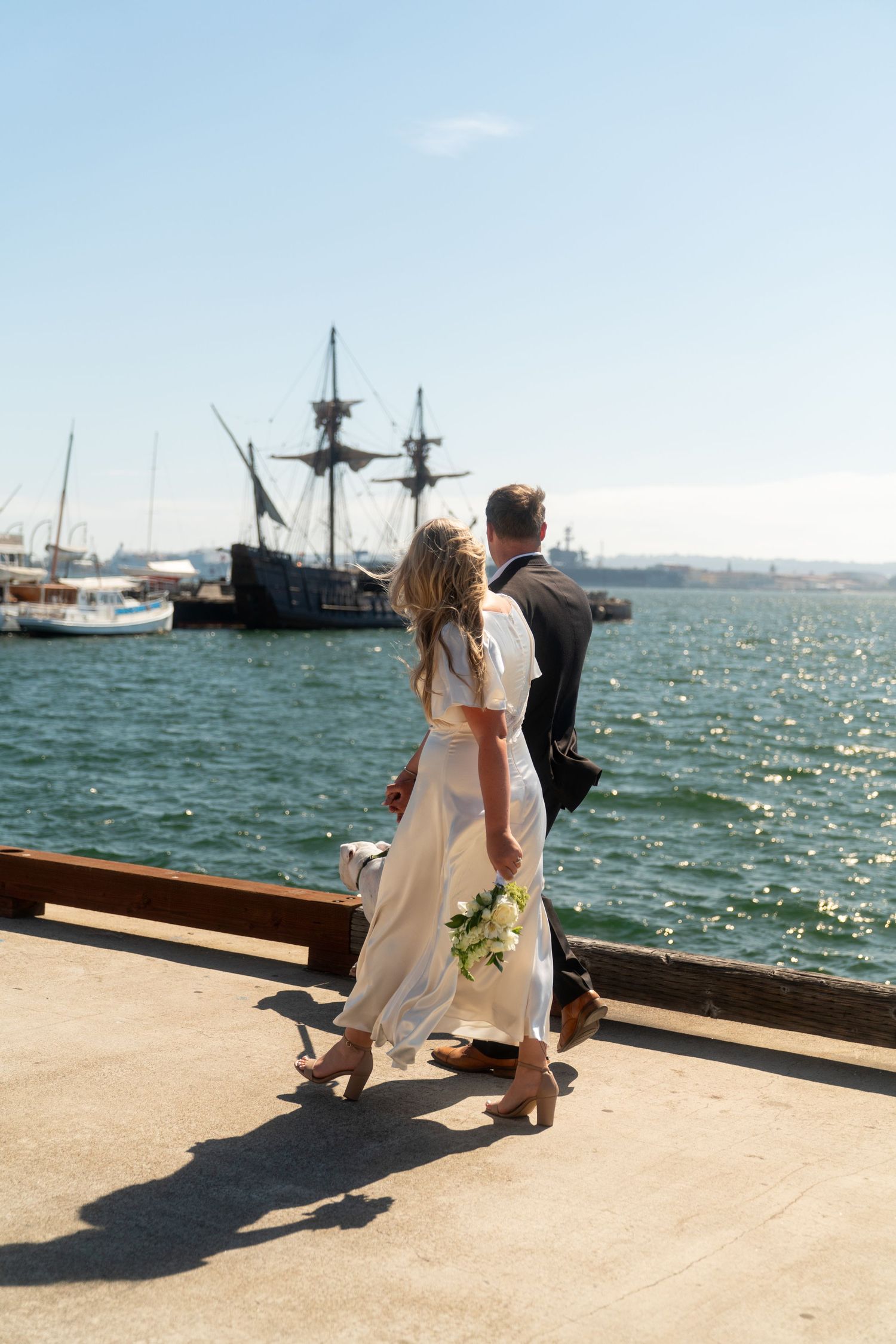 A couple sits on a seaside pier looking out at boats and sparkling water on a sunny day.