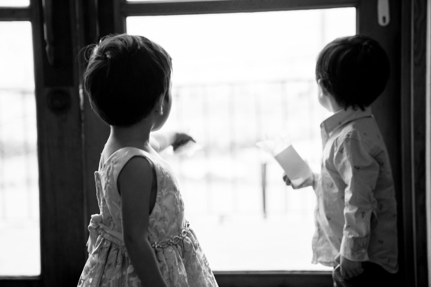 Black and white silhouette of two children looking out window together.