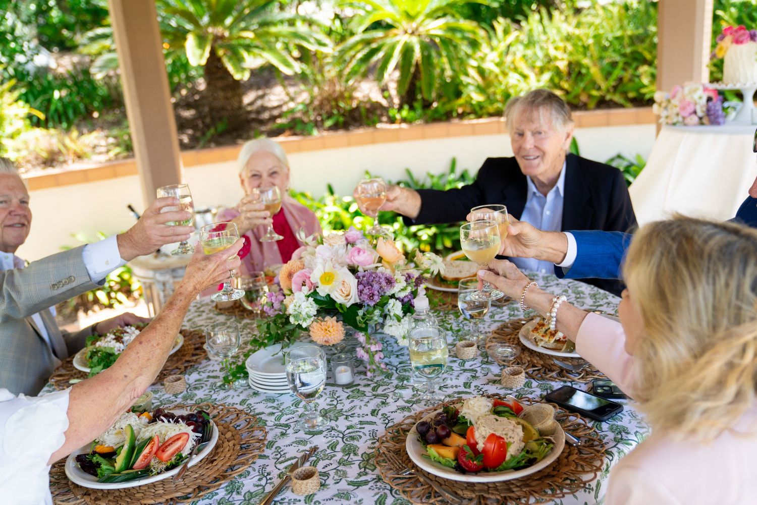 A group gathers for an outdoor meal raising wine glasses in a toast at an elegant table set with flowers and fresh food.