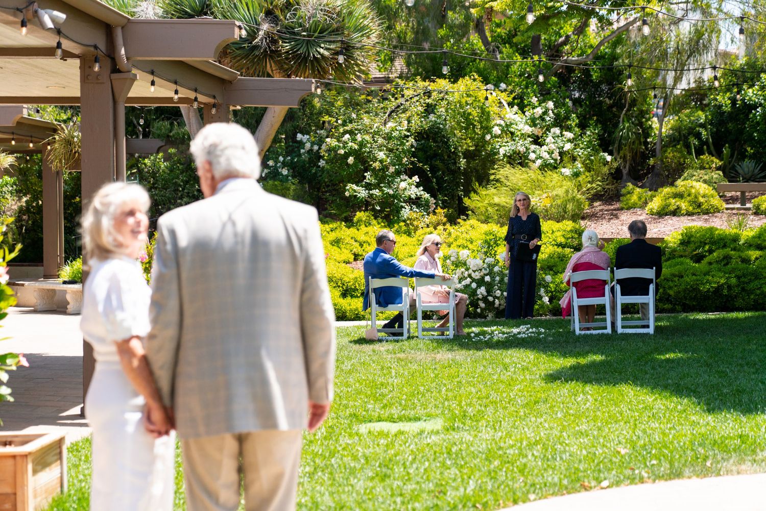 An outdoor wedding ceremony setup in a sunny garden with guests seated on white chairs on the lawn.
