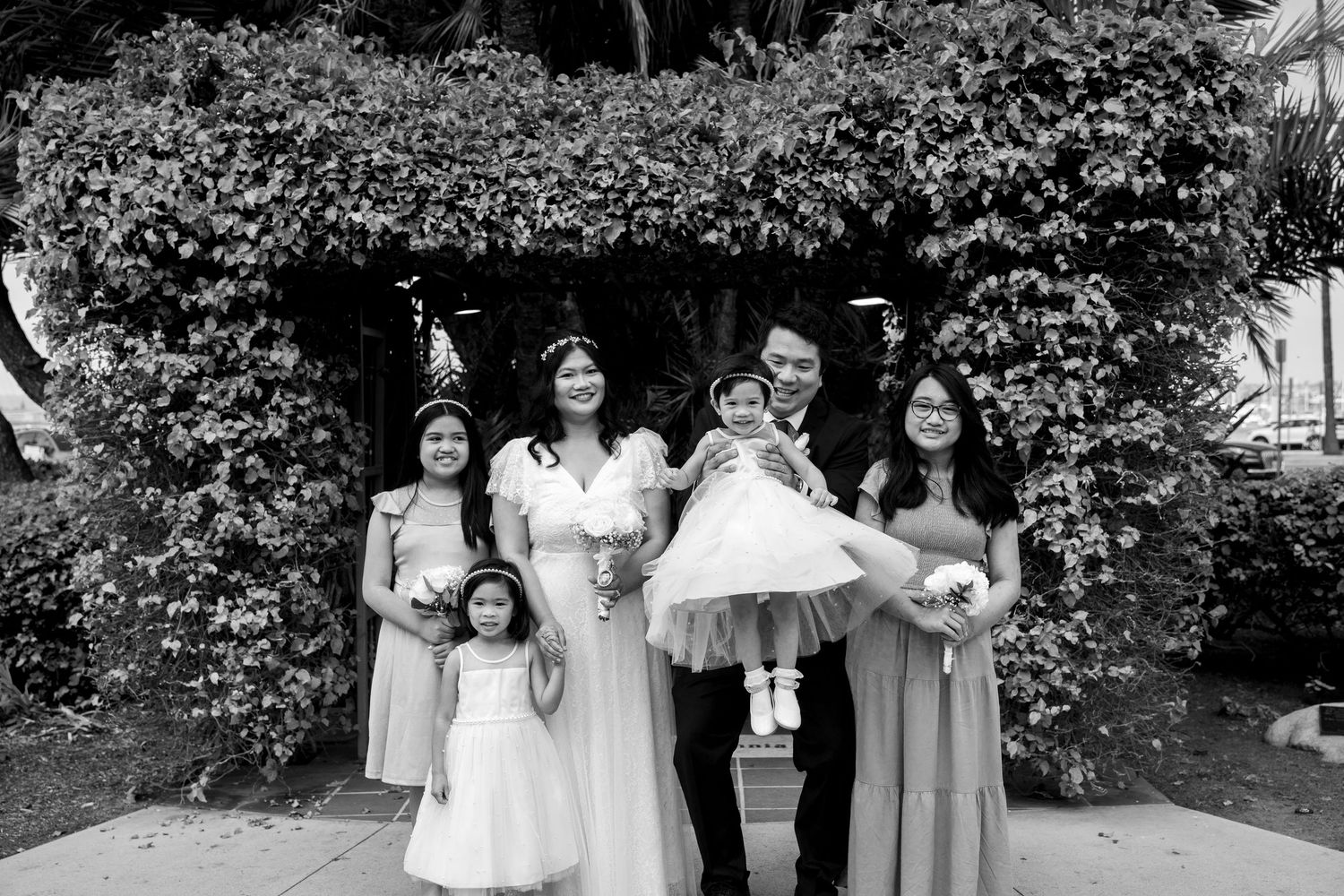 A black and white photo of a wedding party posing outdoors in front of a lush ivy-covered wall.