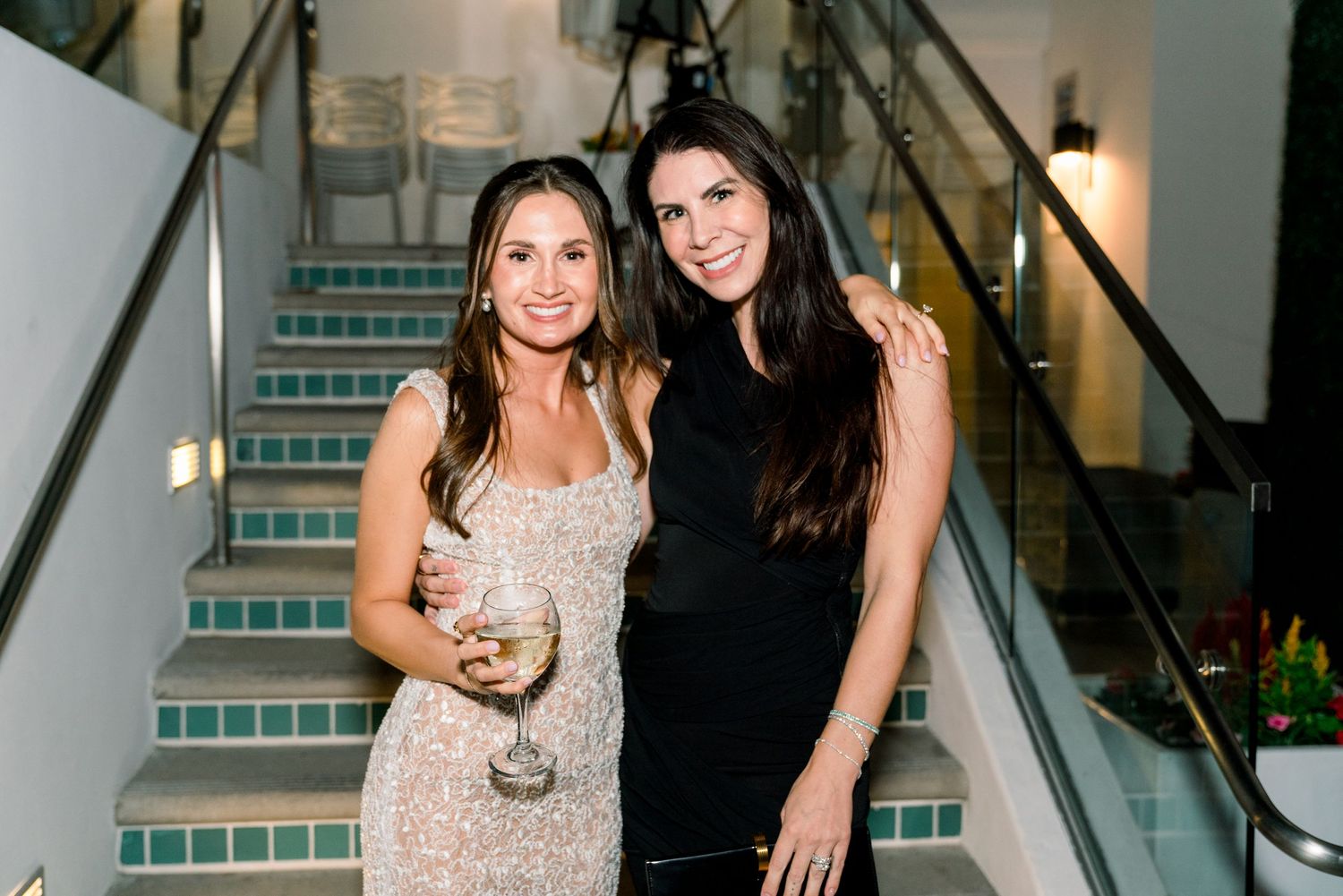 Two friends pose on a staircase at an elegant evening event, one wearing a sparkly dress and one in black.