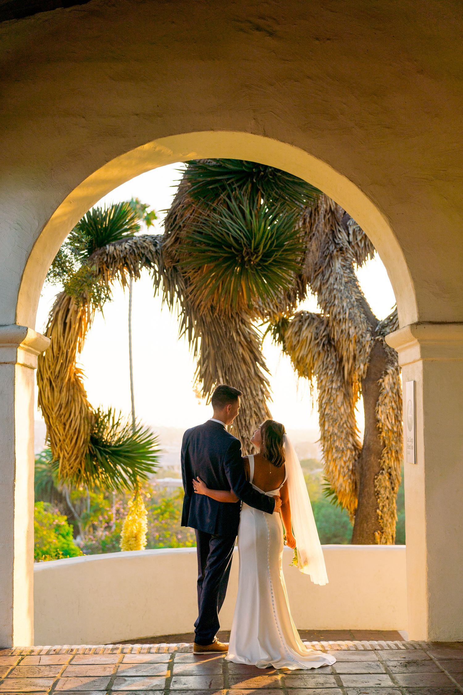 A couple in wedding attire standing under an arched doorway with palm trees in the background.