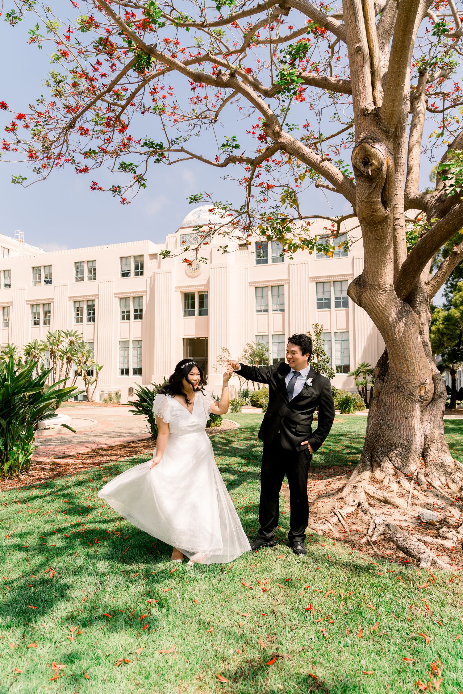 Wedding couple poses under flowering tree in front of vintage building.
