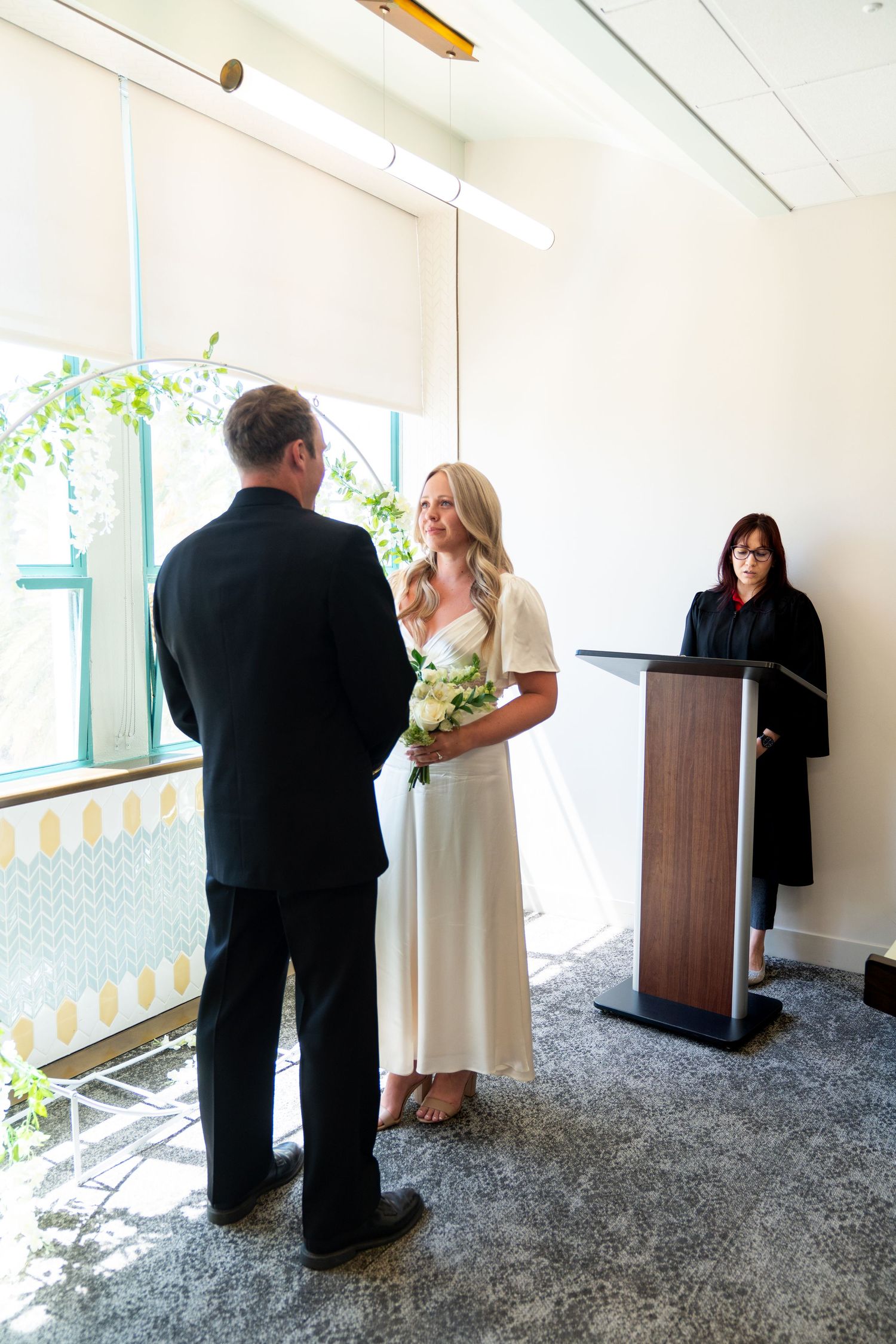 An intimate wedding ceremony takes place in a bright modern room with an officiant at a podium and hanging greenery.