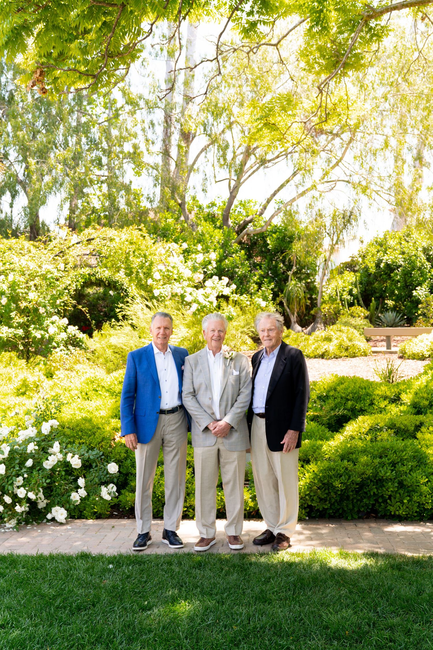 A formal group photo of three people standing together in a beautifully landscaped garden on a sunny day.