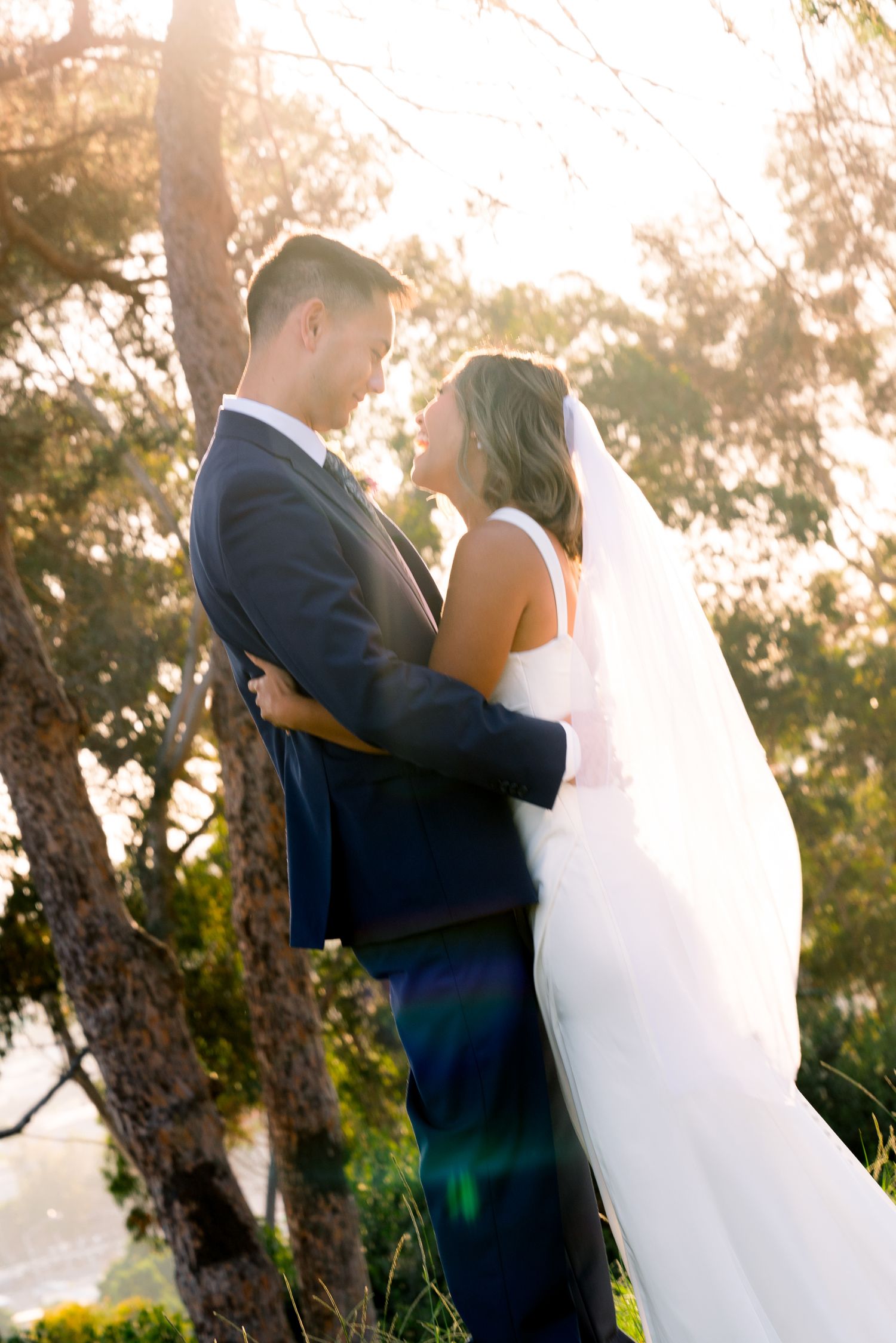 A romantic sunset wedding portrait captures a couple embracing outdoors with sunlit trees in the background.