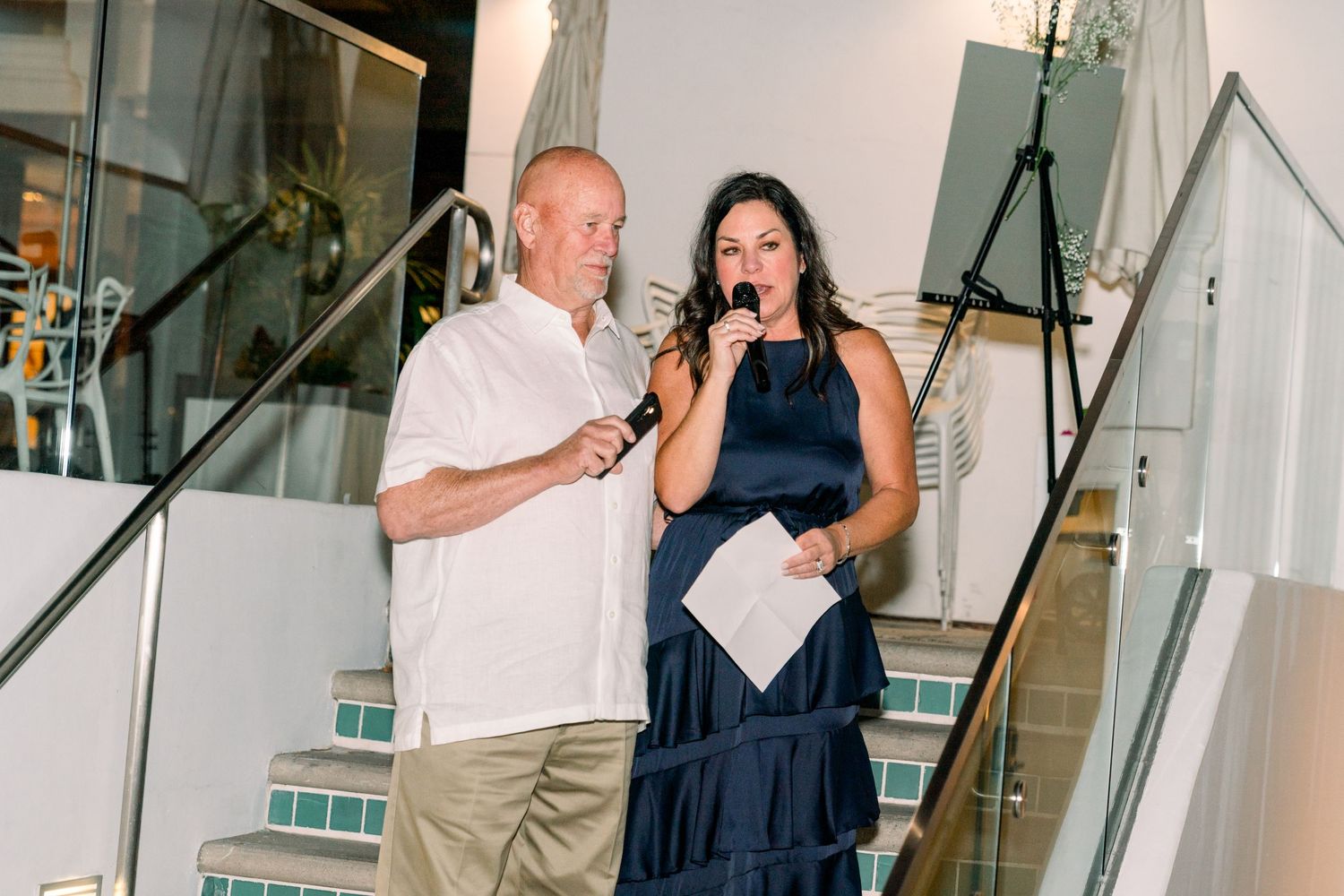 Two speakers share microphones while standing on illuminated stairs during an evening event.