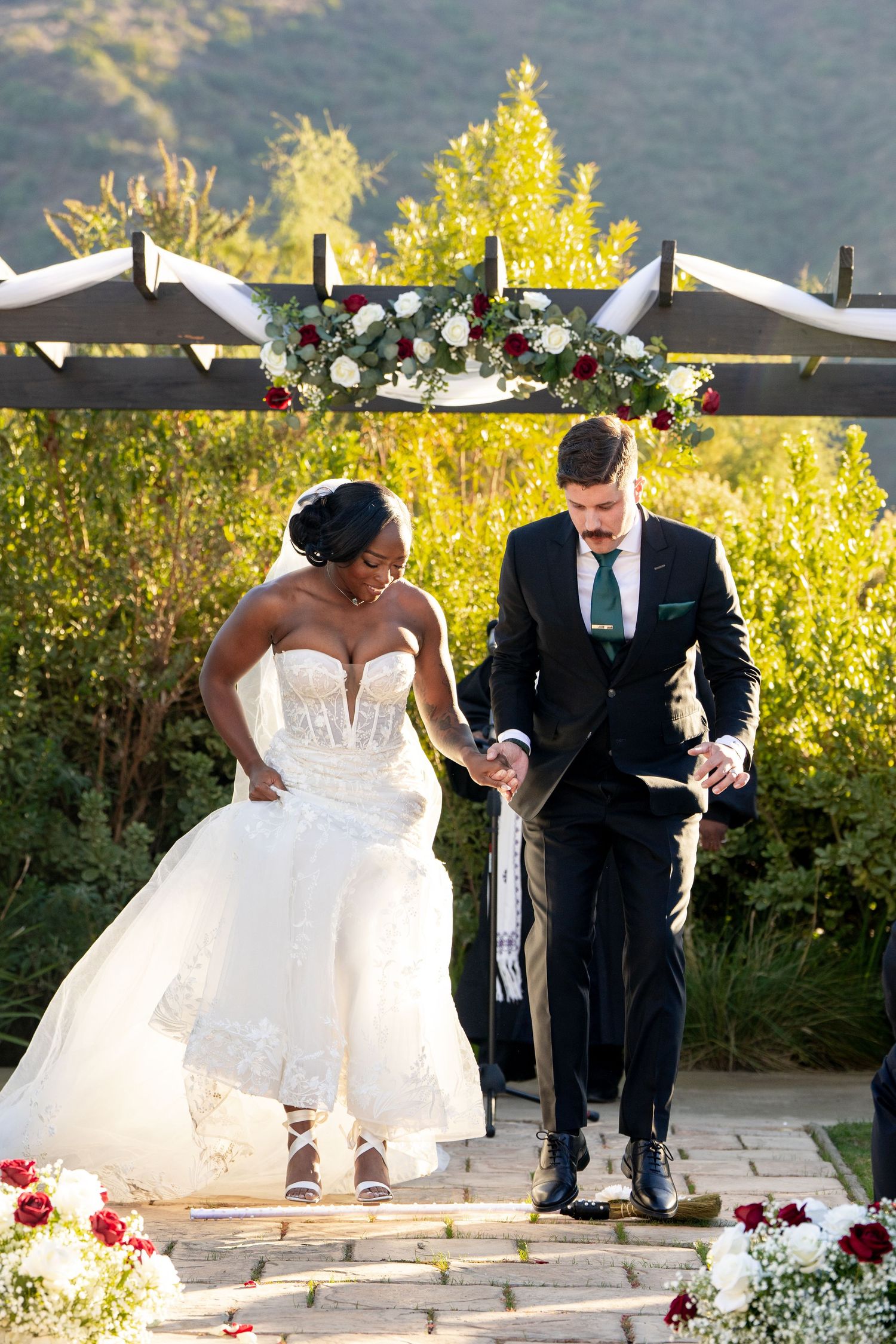 Newlyweds performing unity ceremony under floral arch with mountain backdrop.