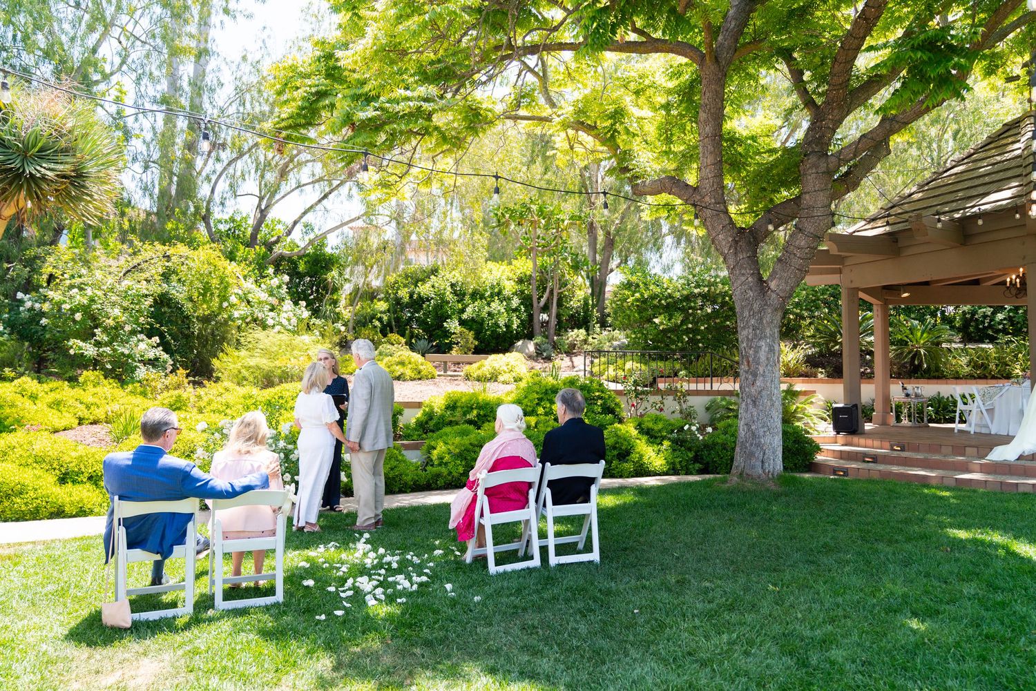 Outdoor garden wedding ceremony setup with white chairs under a shady tree.