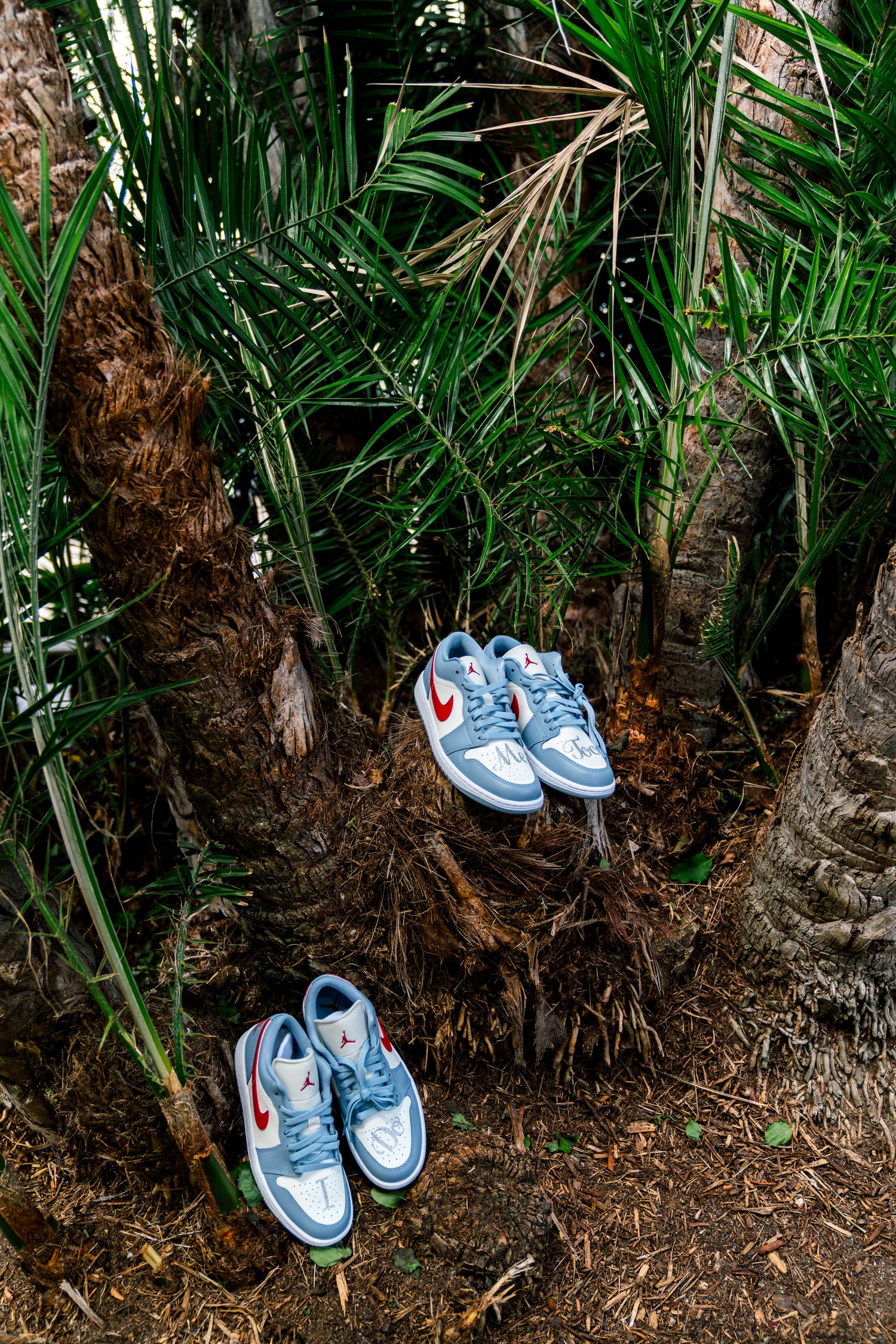 Two pairs of light blue Nike sneakers placed among tropical palm fronds and tree roots in an outdoor setting.