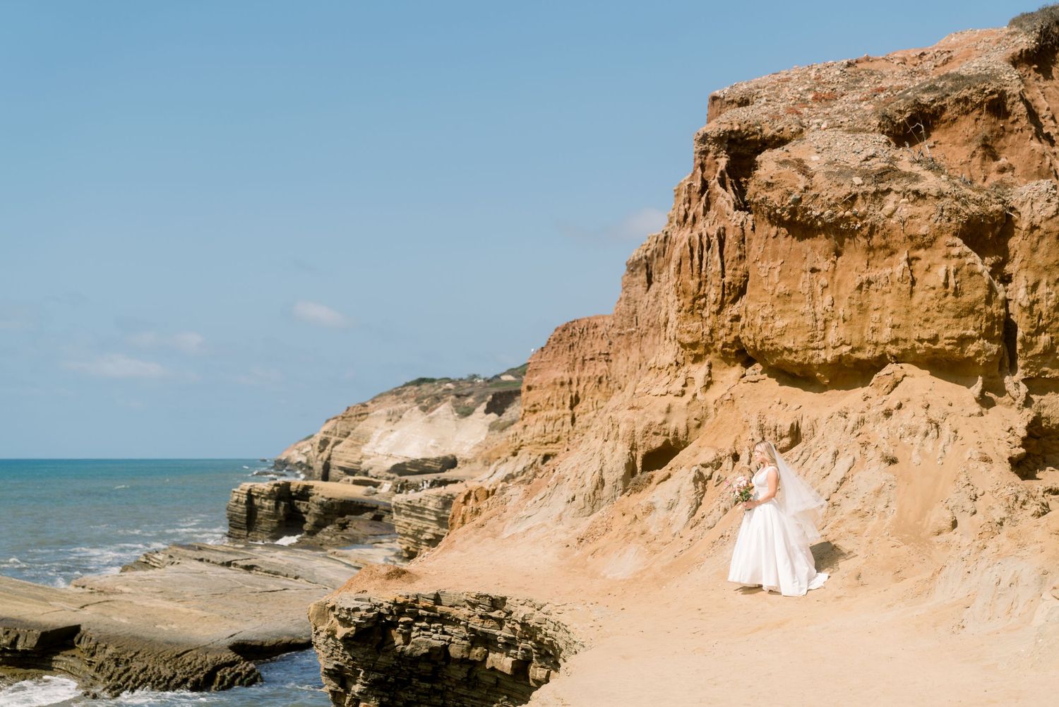 A person in white stands on a sandy beach below towering orange cliffs beside the ocean on a sunny day.