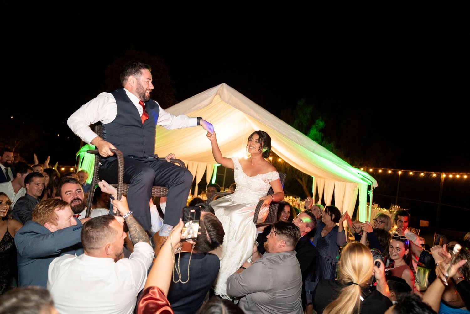 A celebration at a wedding reception with guests lifting the couple on chairs under a tent at night.