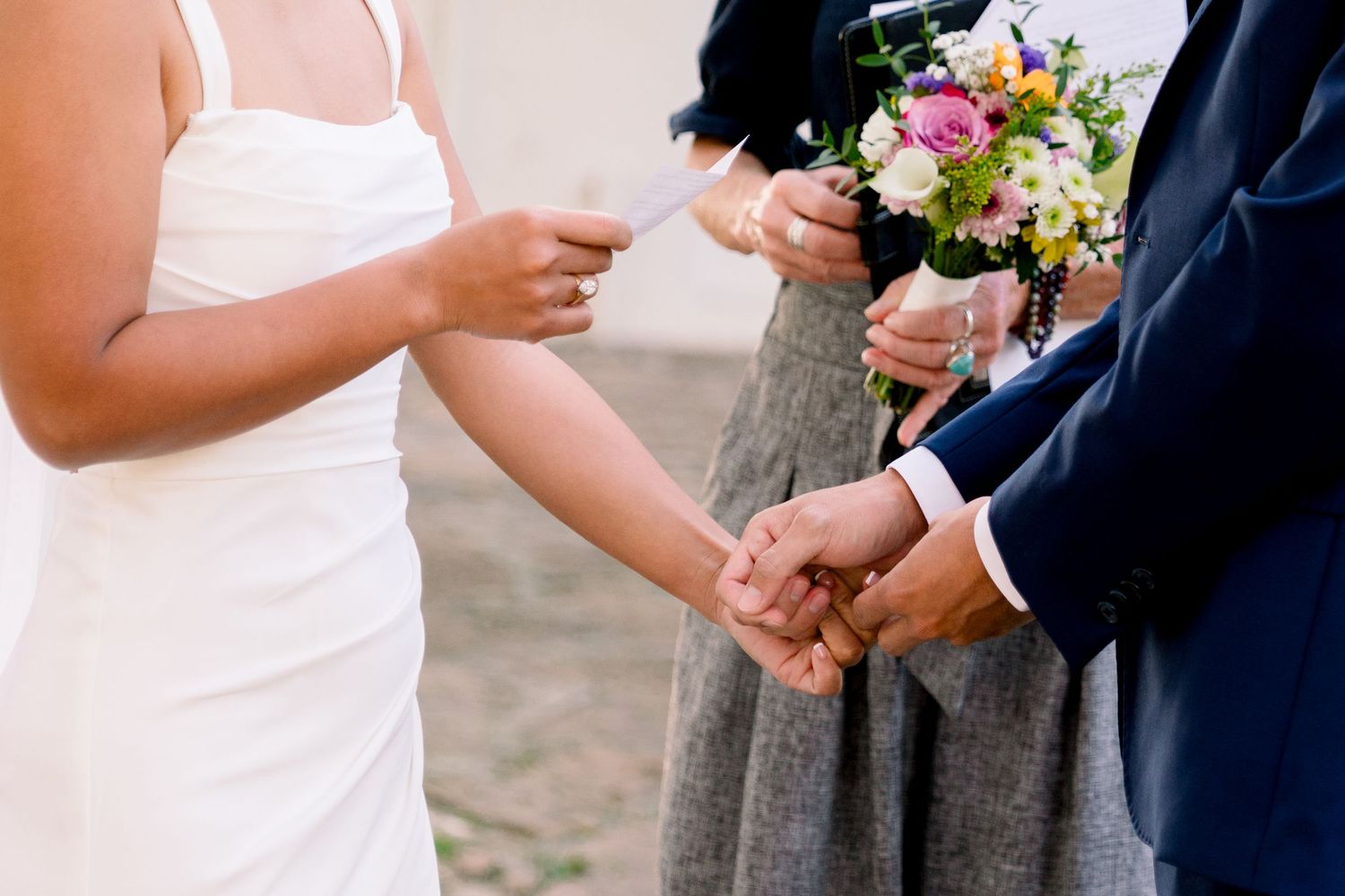 A couple holds hands during an outdoor wedding ceremony with pink and white floral bouquet.