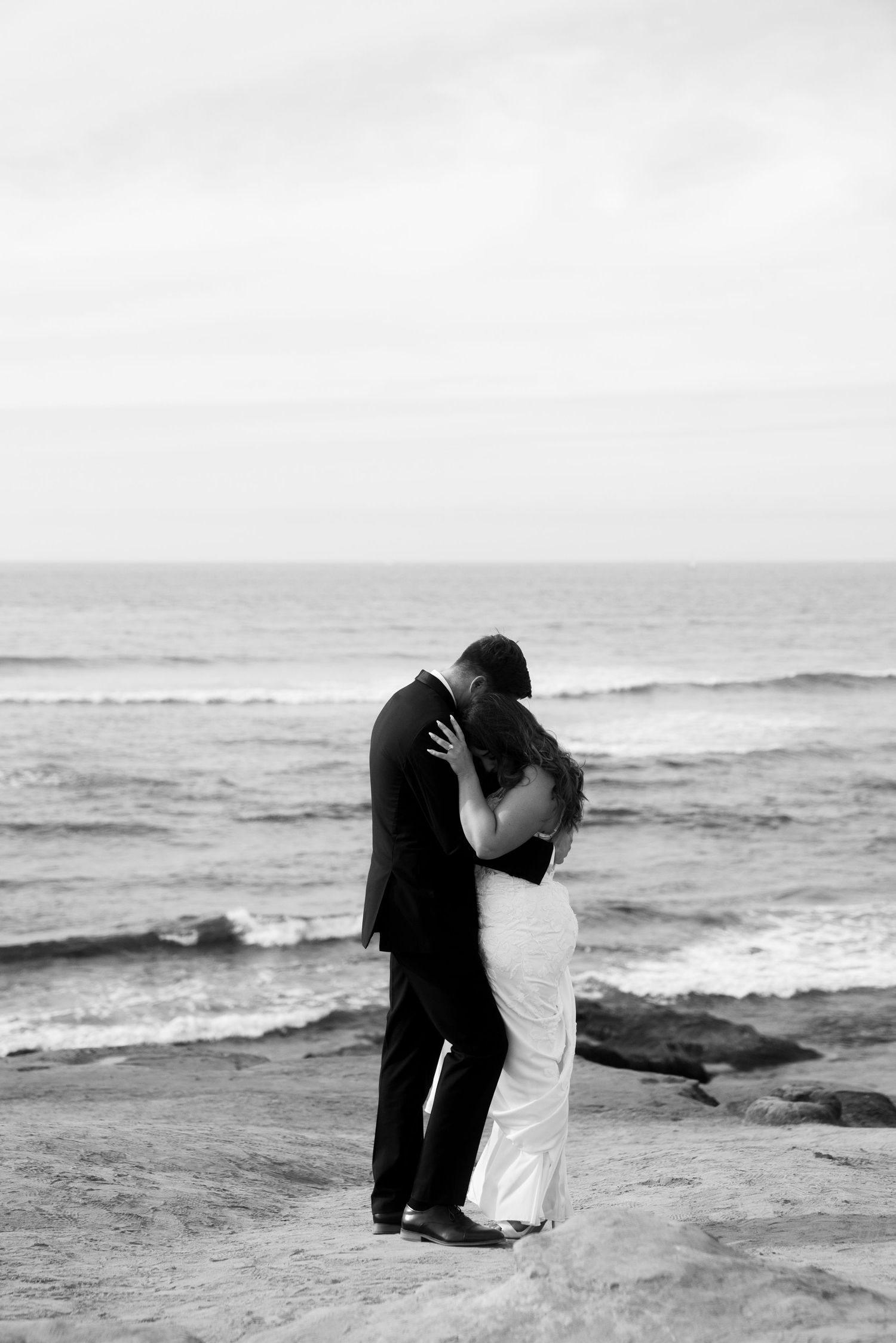 A romantic black and white photo of a wedding couple embracing on rocky coastal cliffs overlooking the ocean.