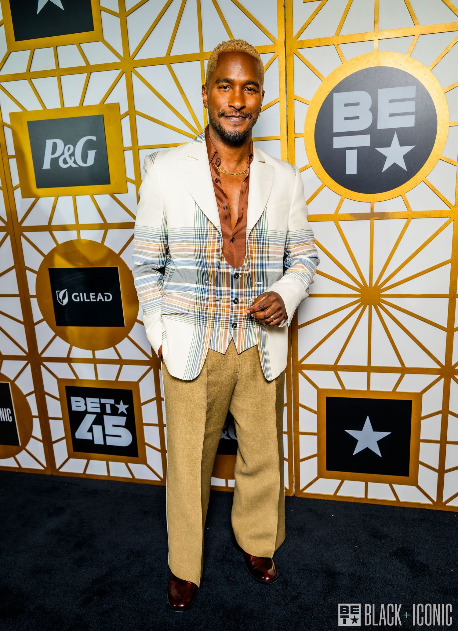 A person poses against a geometric gold and black backdrop at an awards show red carpet event.