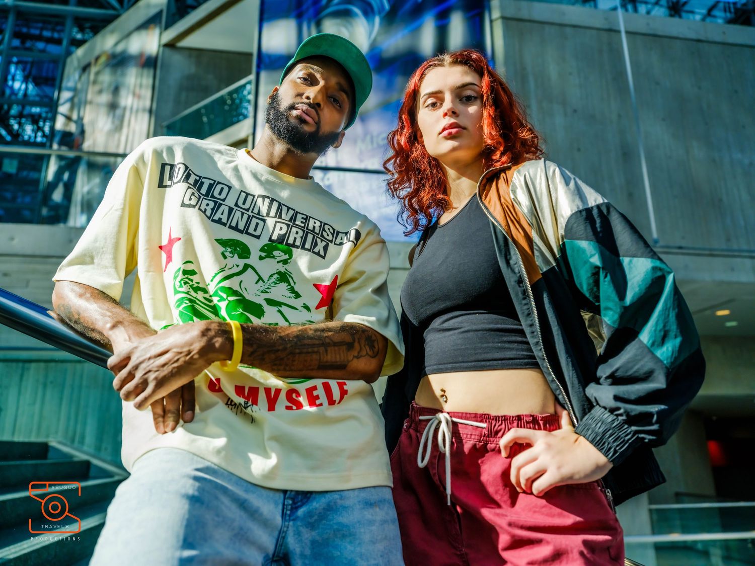 A couple in casual clothing poses playfully against a glass building background in an urban setting.