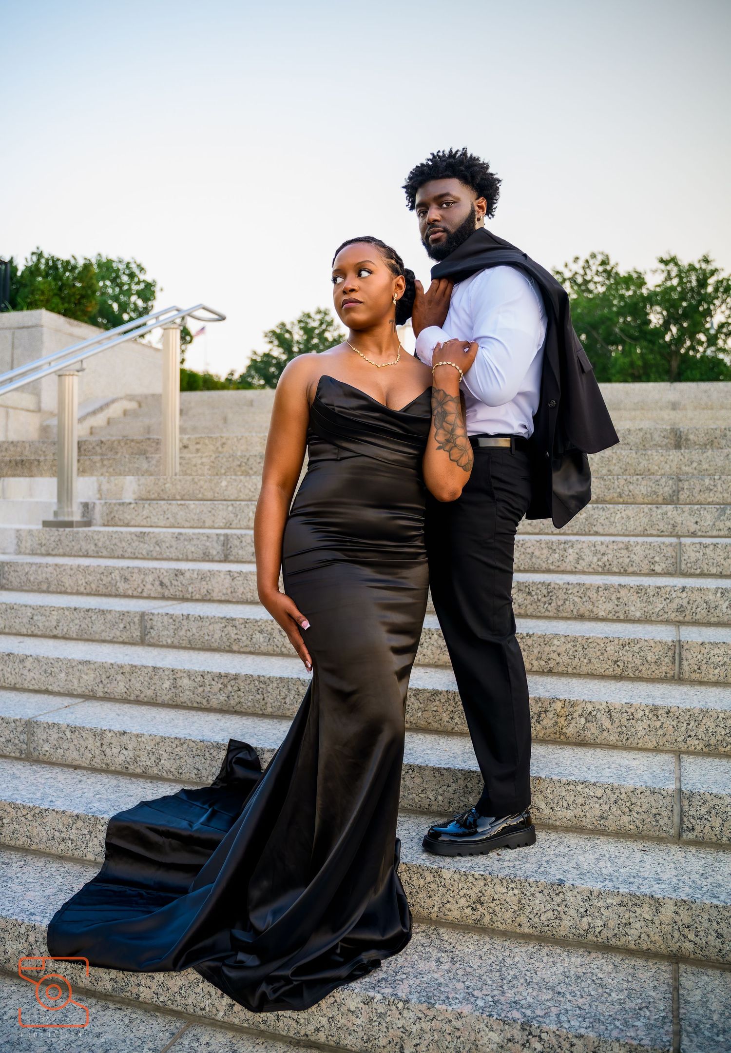 Couple posing on stone steps, woman in elegant black satin gown, man in formal black suit holding jacket.