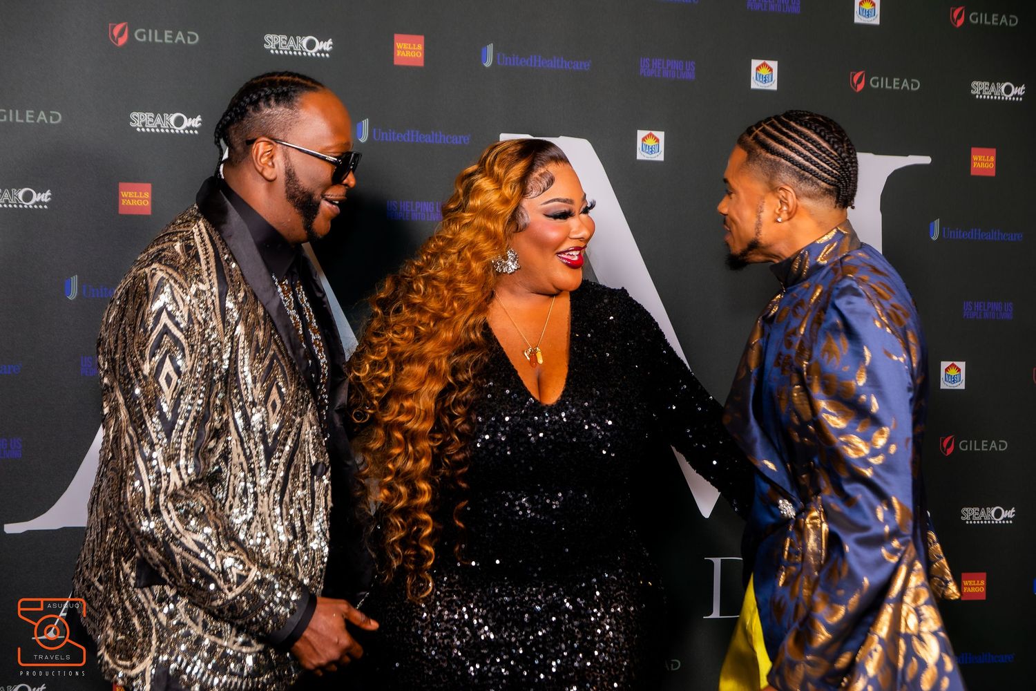 Three performers in glamorous sequined outfits share a moment on a red carpet at an awards show media wall.