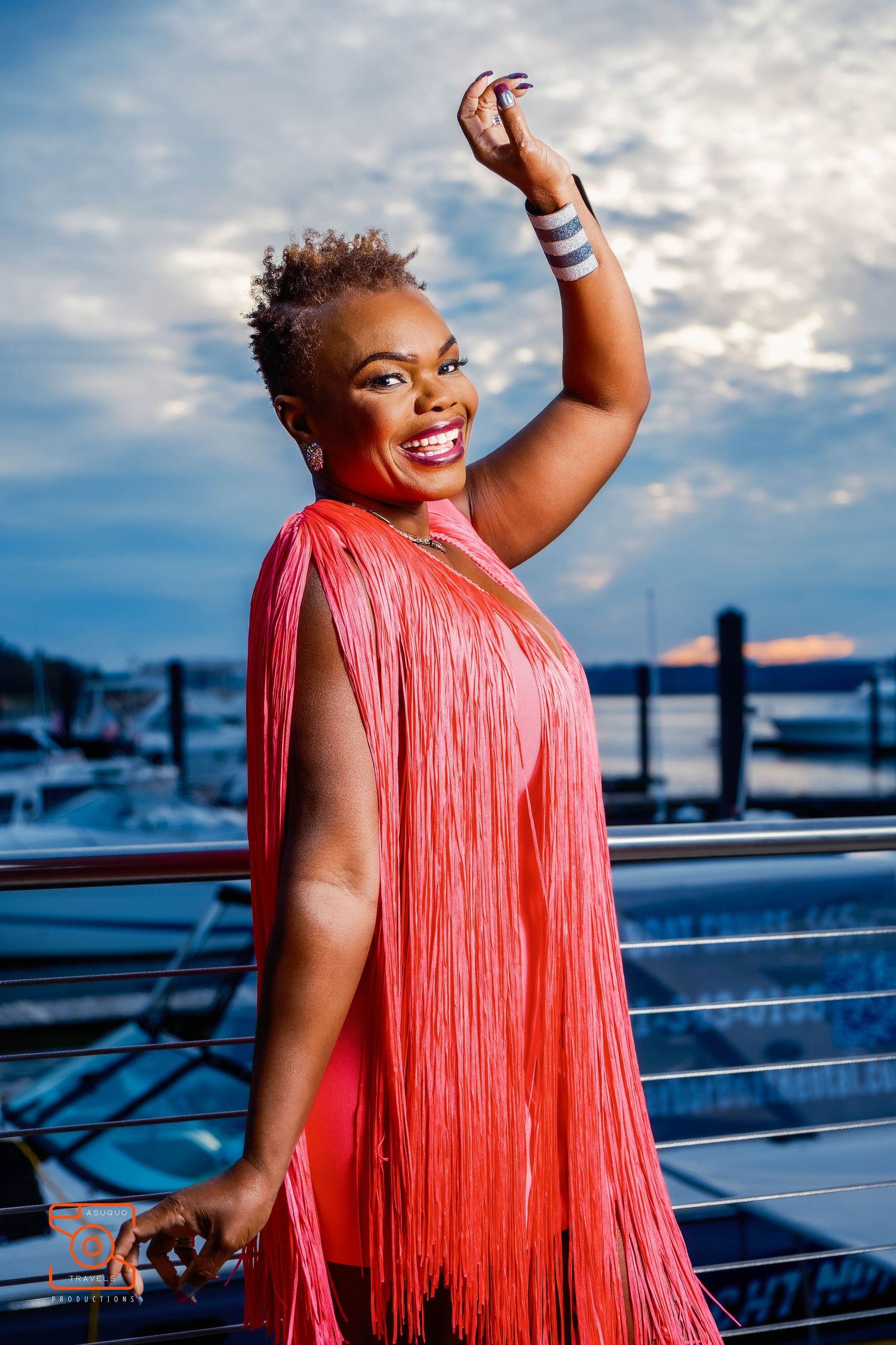 A person in a bright coral fringe dress poses joyfully against a blue sky and waterfront backdrop.