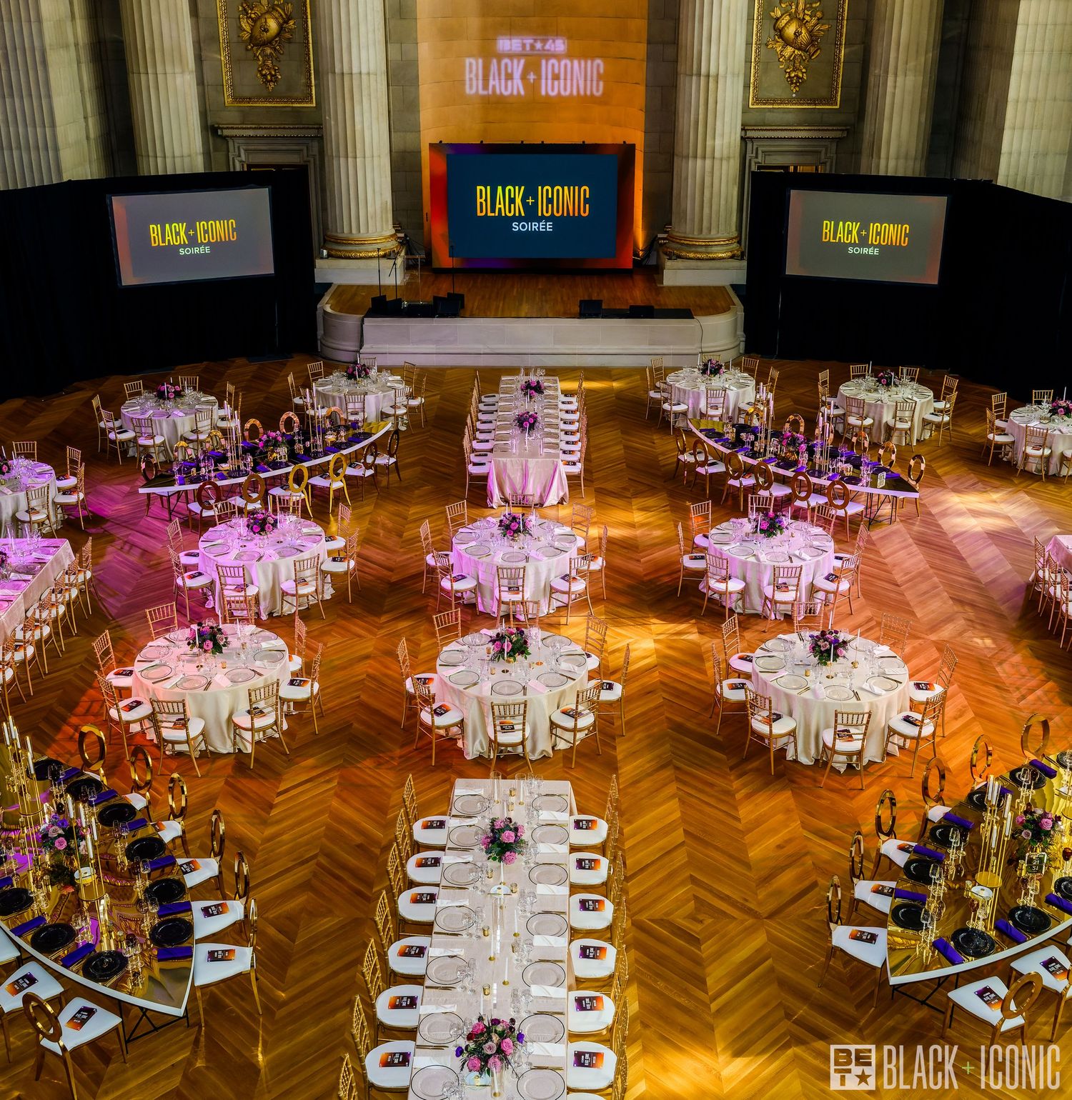 Panoramic view of an elegant ballroom with herringbone wood floors and round dining tables arranged under crystal chandeliers.