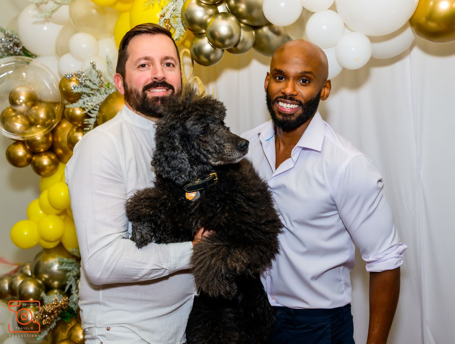 Two people stand together holding a large black fluffy dog in front of a gold and white balloon arch backdrop.