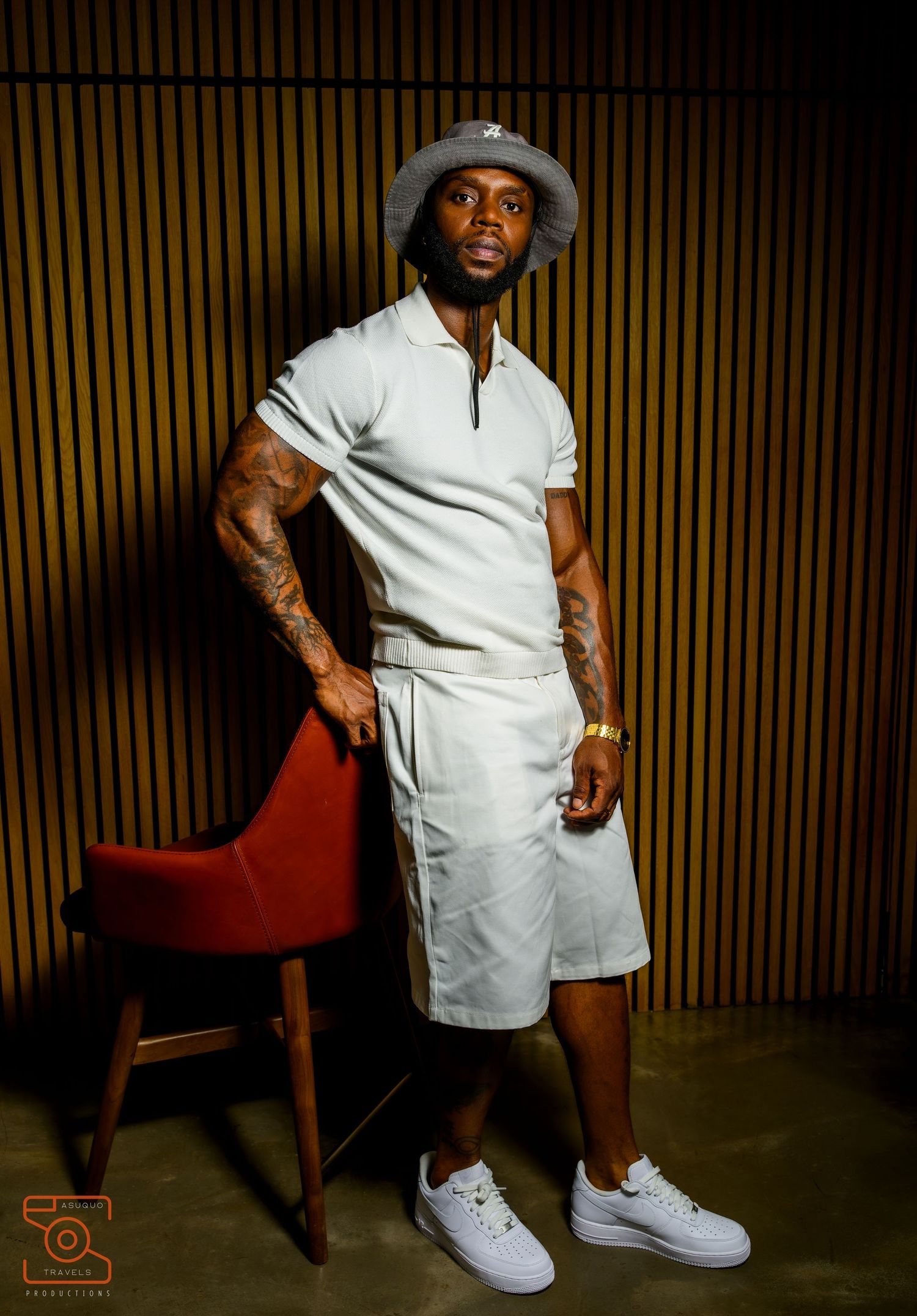 A person in white attire and sneakers poses against a wood-paneled wall with dramatic lighting and a red chair.