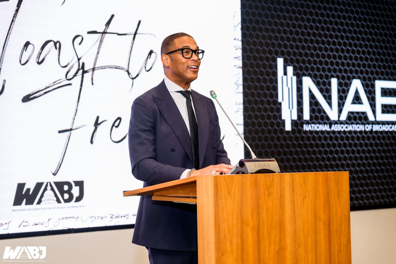 A man in a suit speaks at a podium during a NABJ and WABJ event, with logos visible in the background.