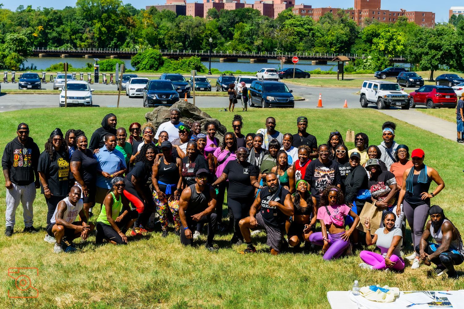 Large group of people sitting together outdoors on grass during a sunny day at a social gathering or event.
