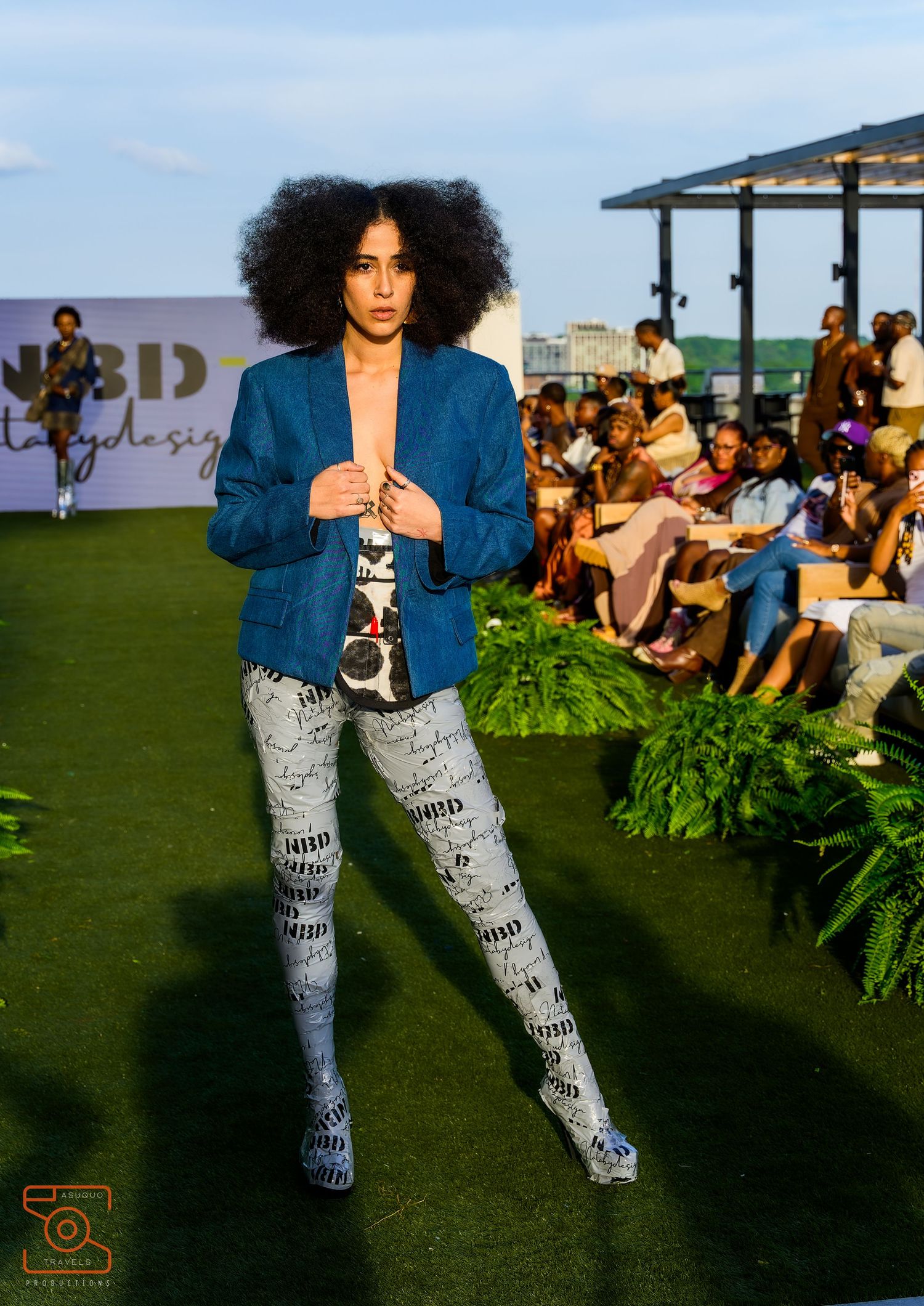Model walks runway wearing blue denim jacket and patterned leggings at outdoor fashion show event.