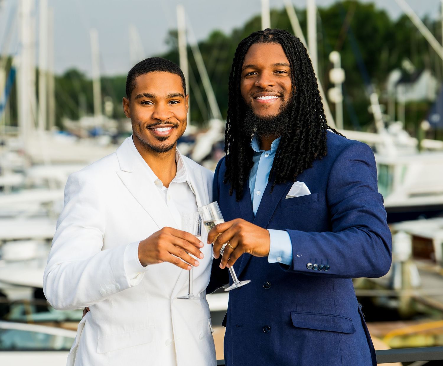 Two people in matching navy and white formal attire smile and embrace in front of a marina setting.