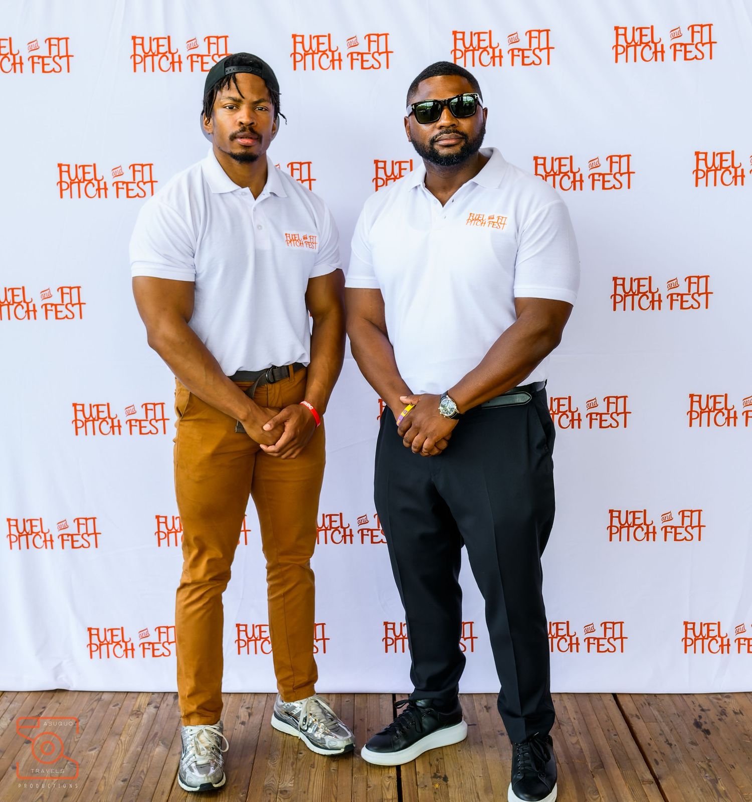 Two people wearing white polo shirts and brown pants stand against a wall with branded event signage at a festival.