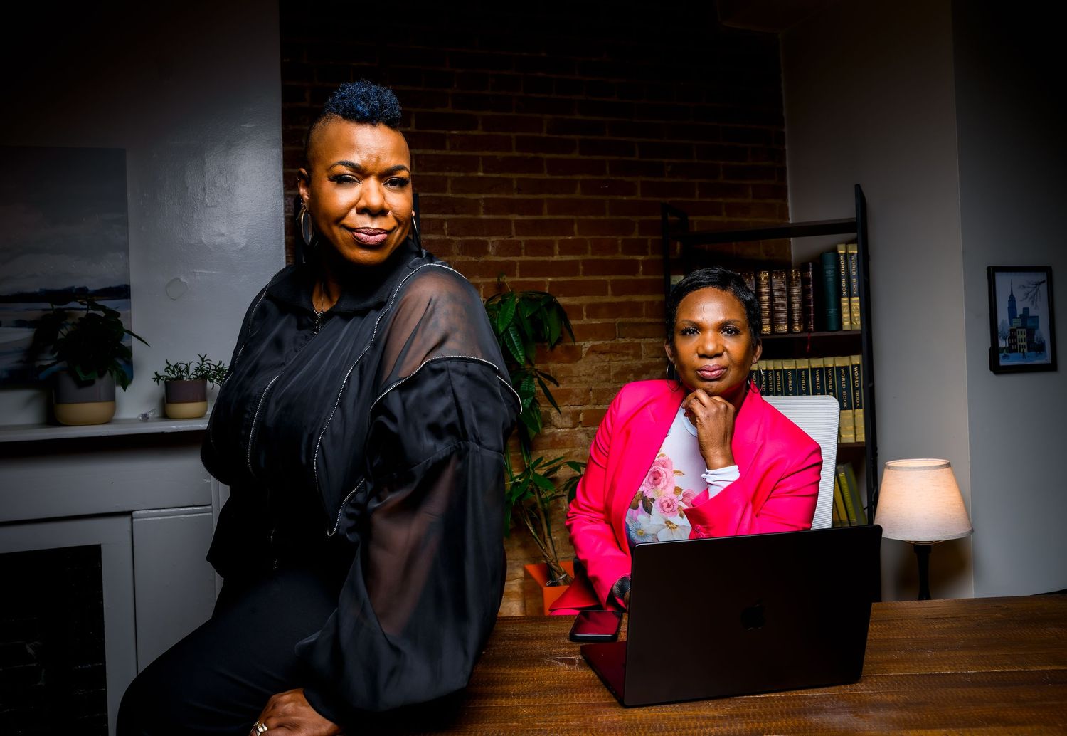 Two professional women pose in a modern office setting, one standing in black outfit, other seated at laptop in pink blazer.