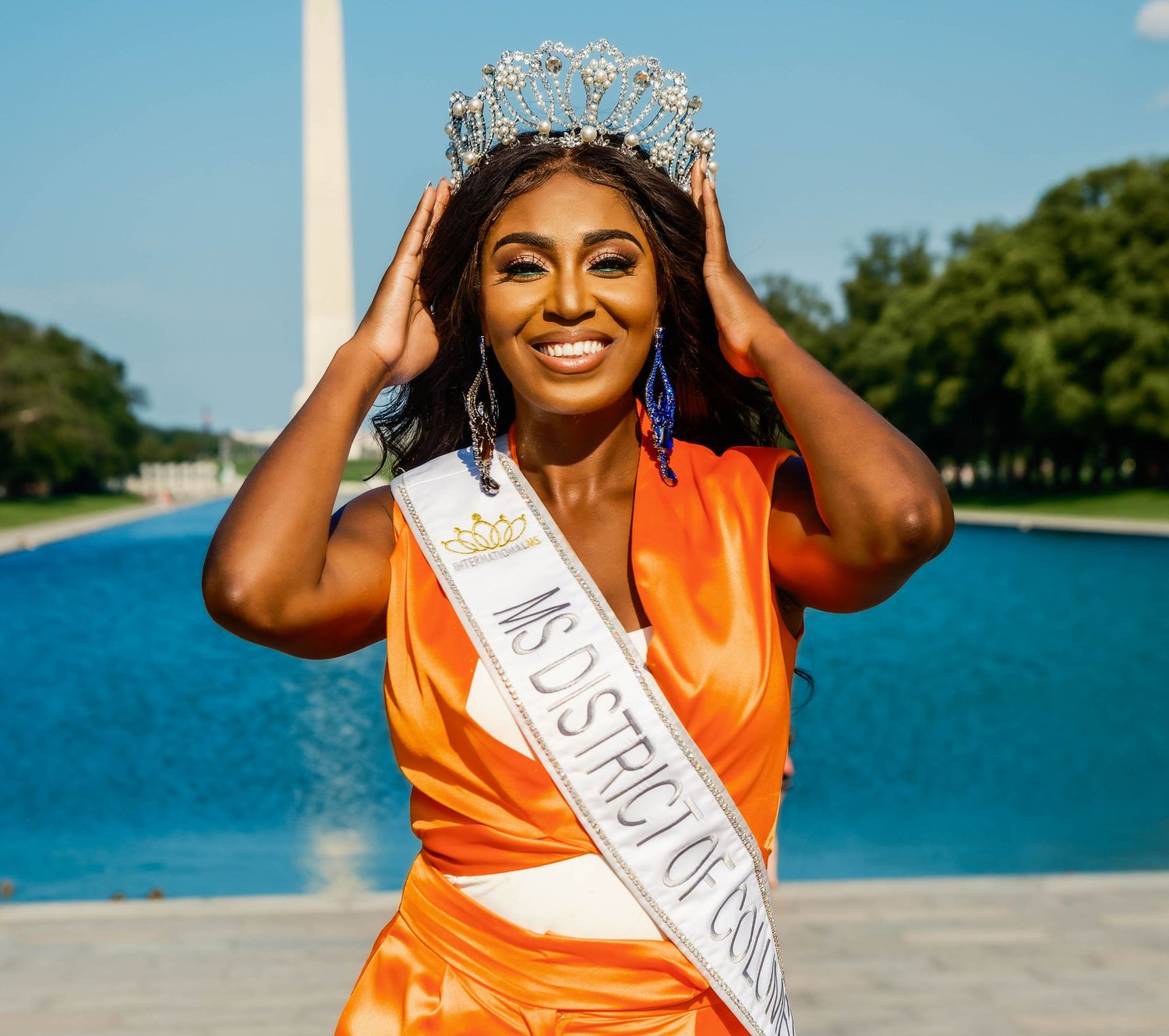 Person wearing orange satin dress and pageant crown poses by waterfront on sunny day.