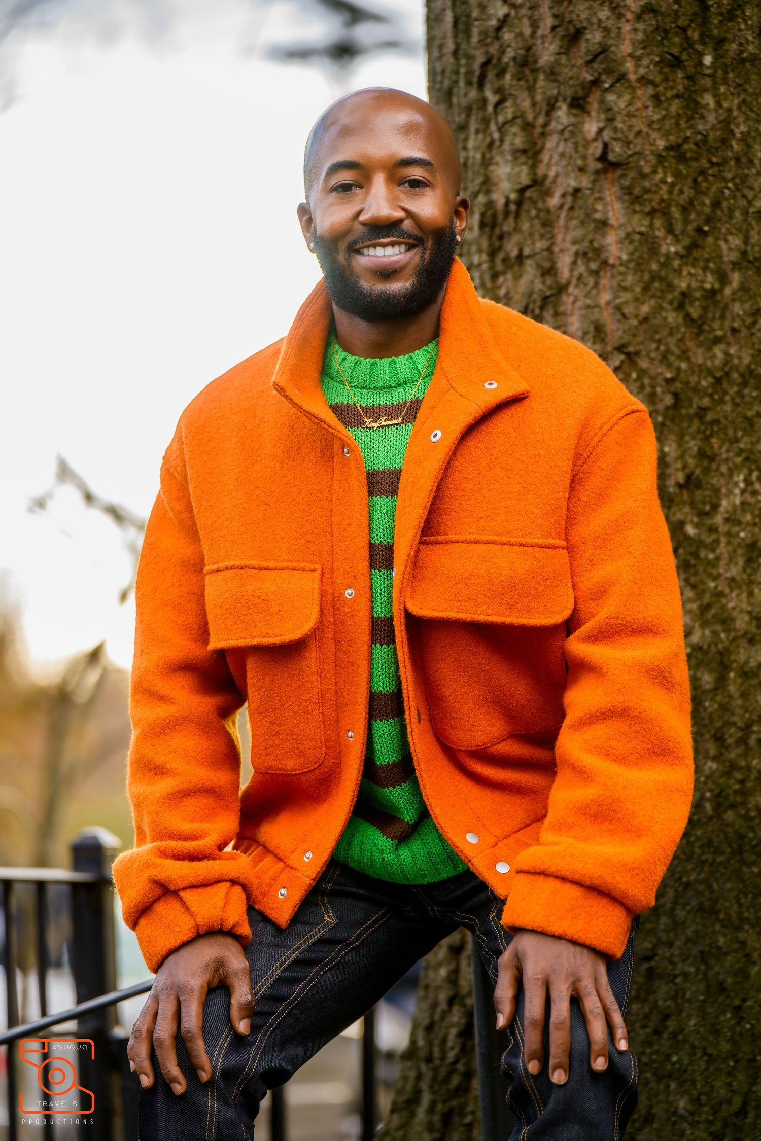 Person wearing bright orange jacket and green sweater sits by tree trunk smiling warmly in outdoor setting.