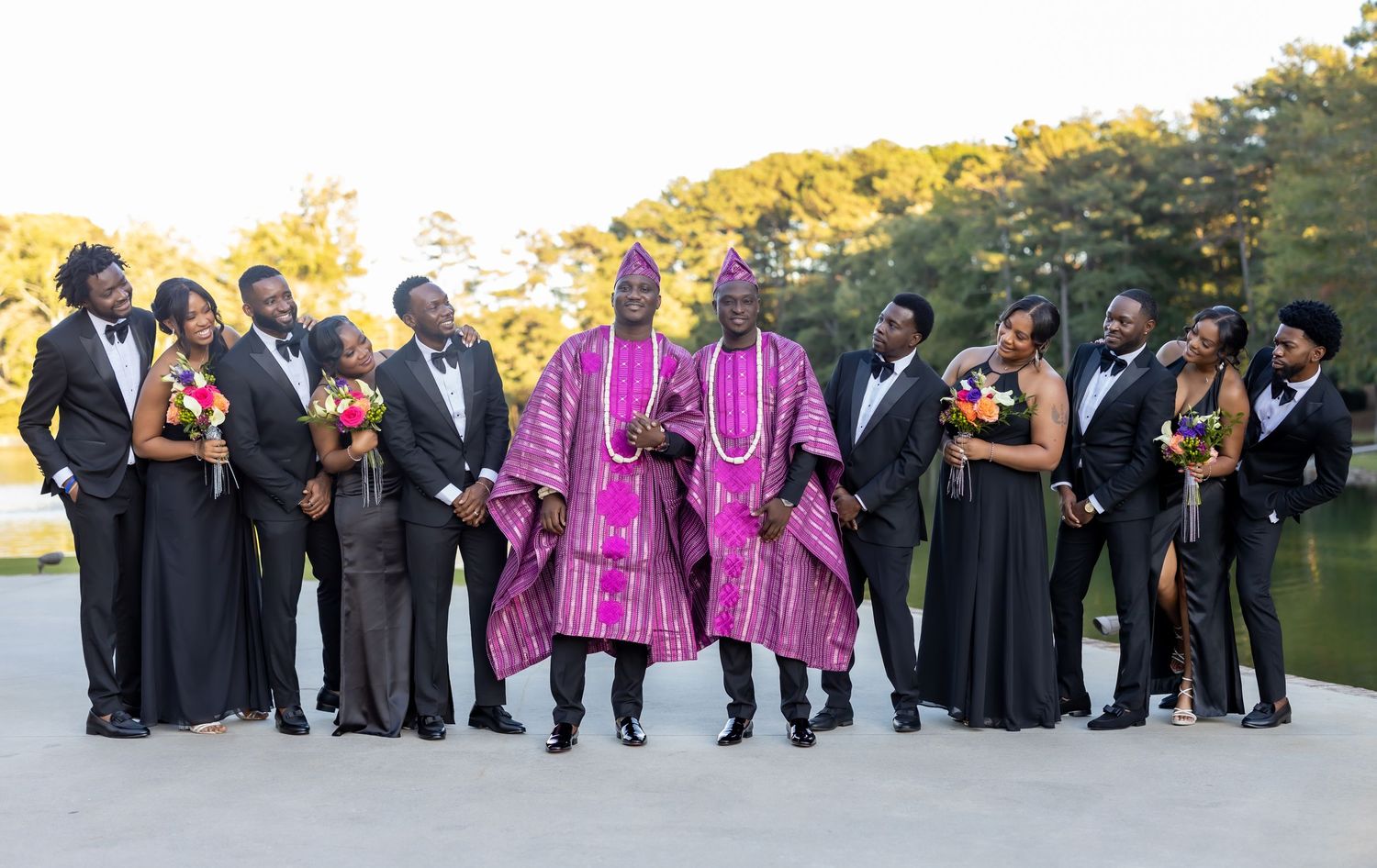 A wedding party in dark formal attire poses with bridesmaids in bright pink dresses outdoors during sunset.