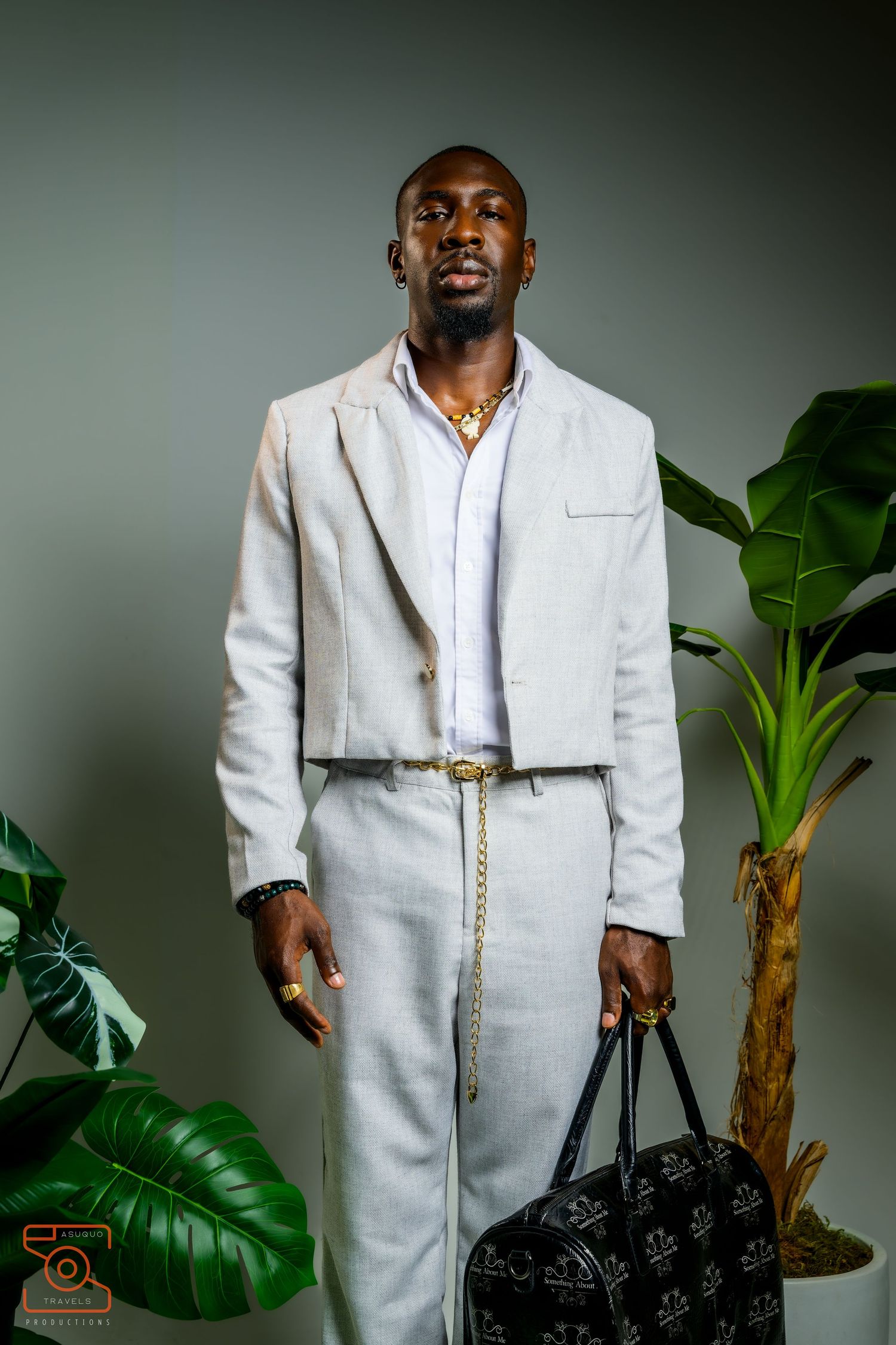 A person wearing a white linen suit and gold chain poses against a gray background with tropical plants.