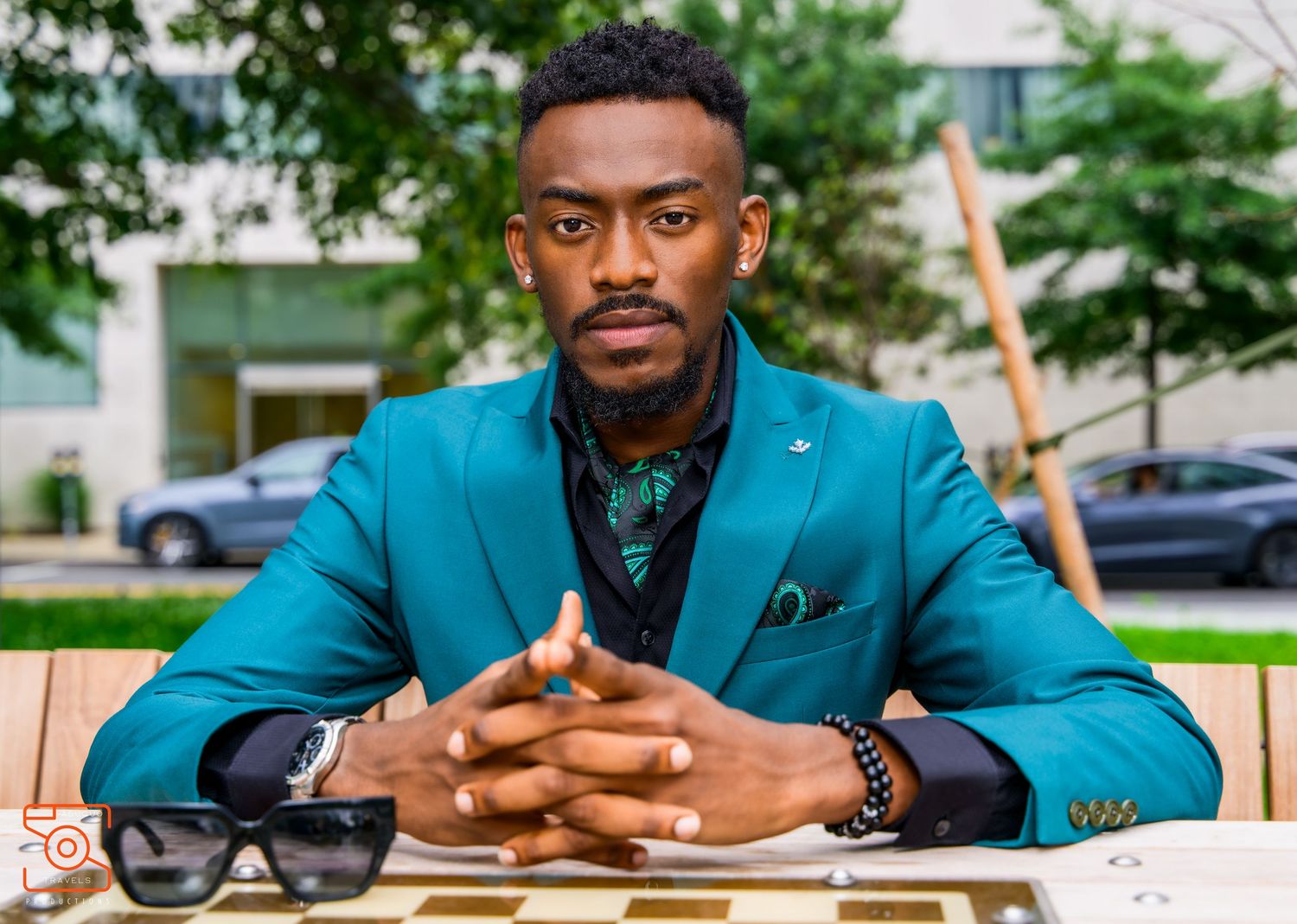 Stylish person in teal blazer and patterned shirt sits at outdoor table with sunglasses and folded hands.