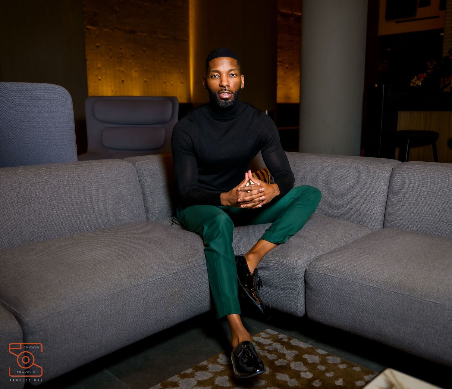 Stylish man in black turtleneck and green trousers sitting confidently on a gray sofa in a modern interior.