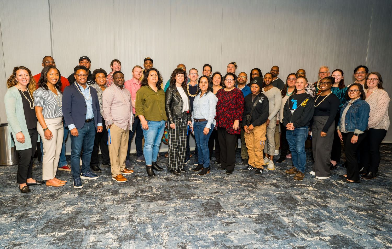 A large group of people standing together for a panoramic group photo against a gray wall background.