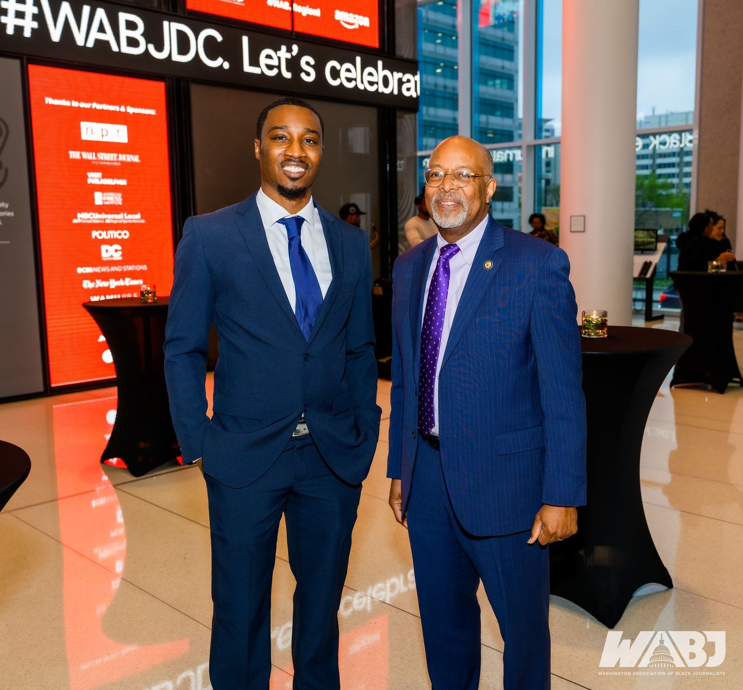 Business professionals in navy suits gathered at a corporate event with red branding displays.